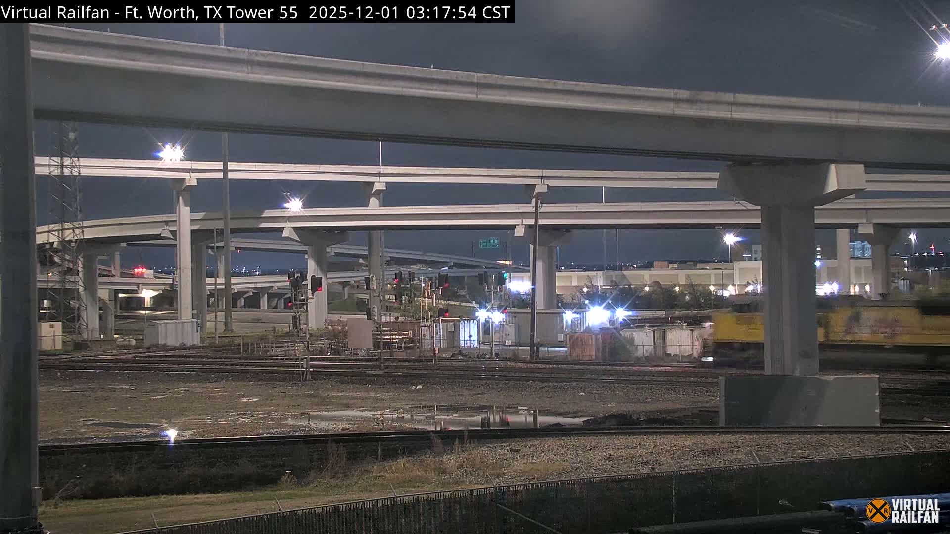 At night, a yellow train moves past a brightly lit, complex elevated highway interchange above wet railroad tracks under a dark sky.