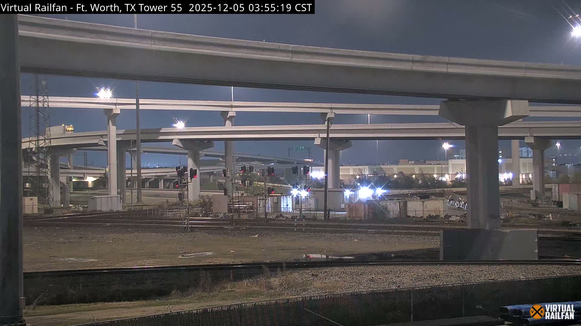 A vast network of concrete highway overpasses looms over multiple railroad tracks and industrial lighting at night, under clear skies.