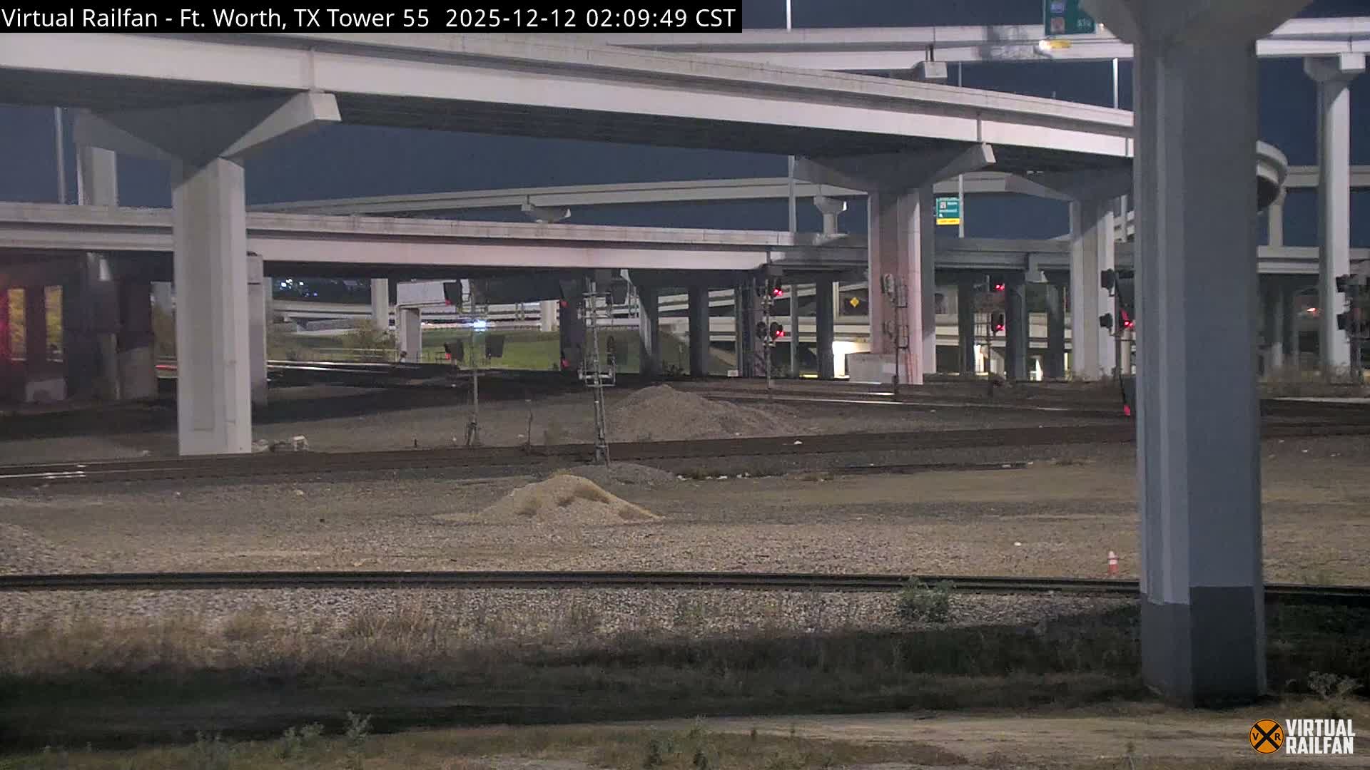 Multiple train tracks traverse the foreground and midground at night beneath a sprawling concrete highway interchange with multiple elevated levels, illuminated by artificial lights and featuring several railroad signals displaying red lights under clear weather conditions.