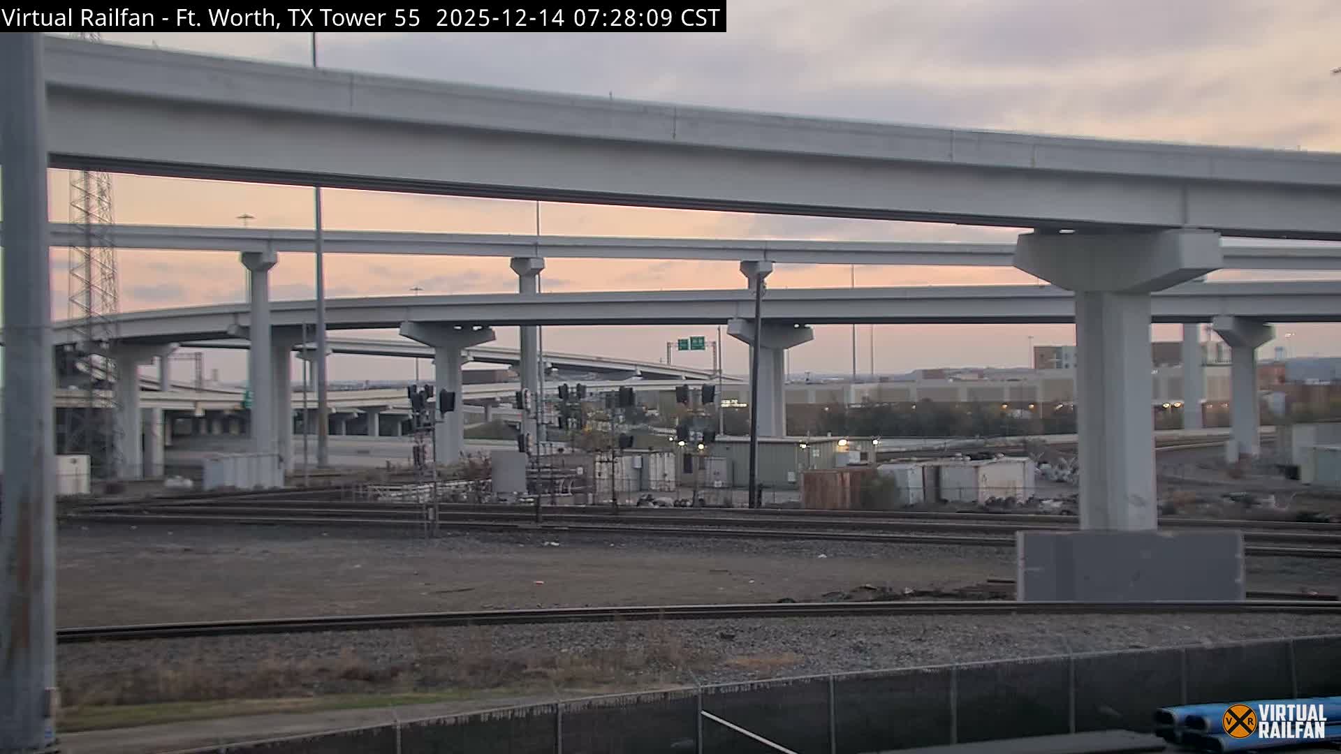 Multiple train tracks traverse the foreground and midground at night beneath a sprawling concrete highway interchange with multiple elevated levels, illuminated by artificial lights and featuring several railroad signals displaying red lights under clear weather conditions.