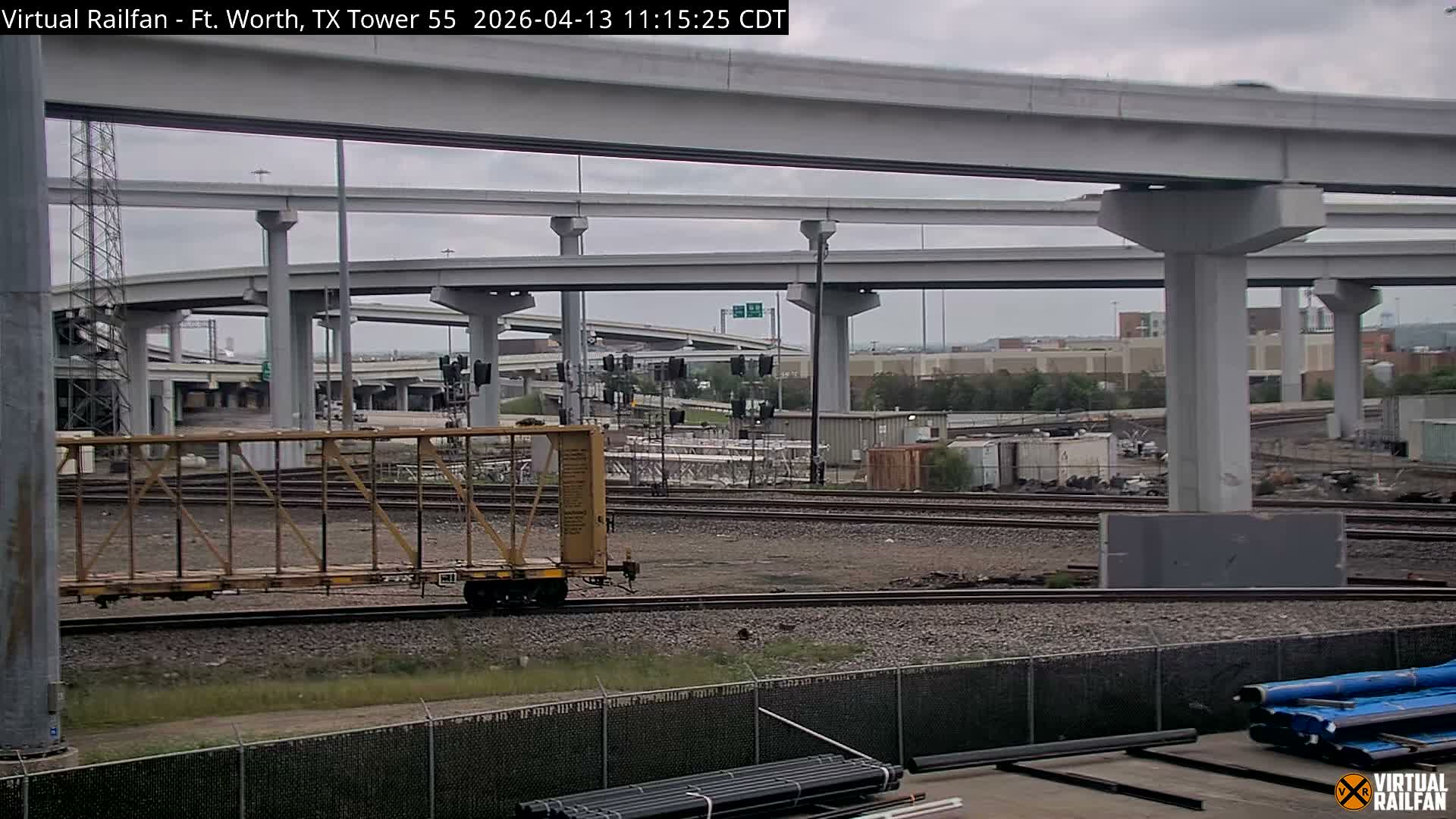 Multiple train tracks traverse the foreground and midground at night beneath a sprawling concrete highway interchange with multiple elevated levels, illuminated by artificial lights and featuring several railroad signals displaying red lights under clear weather conditions.