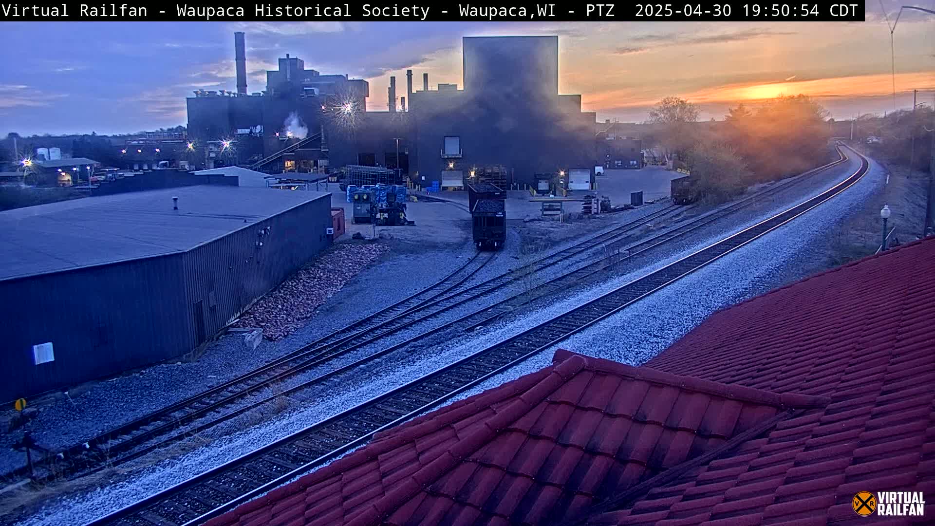 At sunset, a train car approaches a junction near a factory emitting smoke, with a red tiled roof in the foreground.