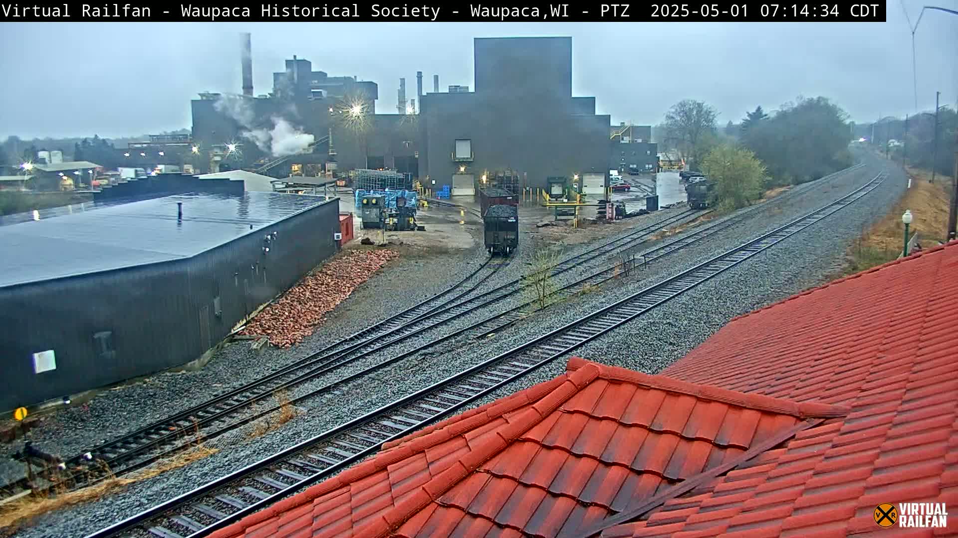 A rain-swept industrial area with railroad tracks, a large factory building emitting smoke, and a red-tiled roof in the foreground.