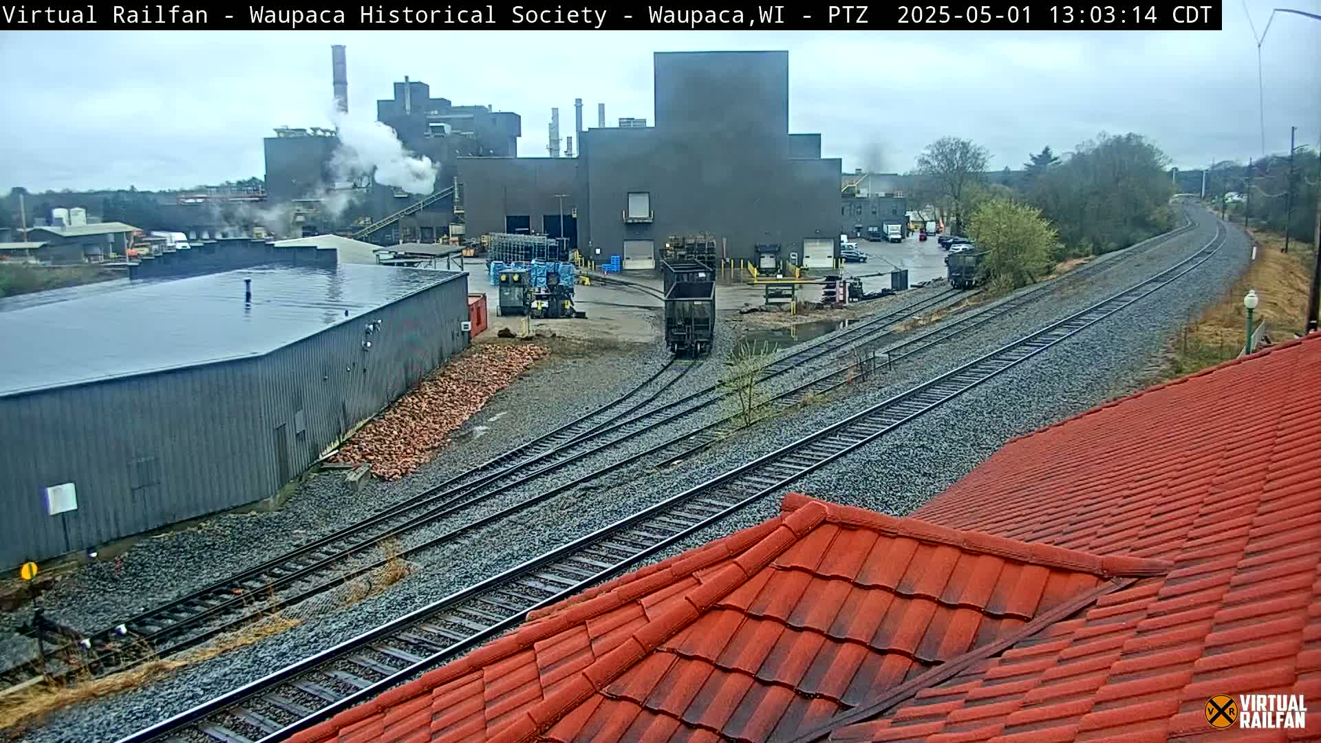 A view of an industrial area with a large factory emitting smoke, railroad tracks, and a red tile roof in overcast conditions.