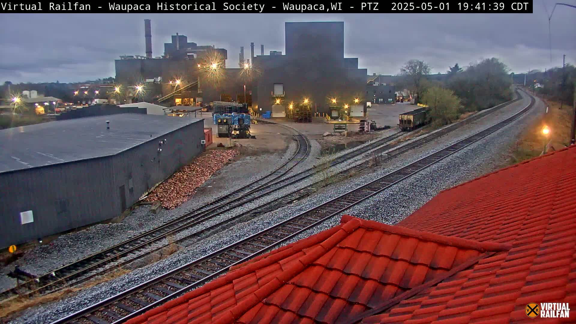 A view of railroad tracks curving past an industrial area under an overcast sky, with a red roof in the foreground.
