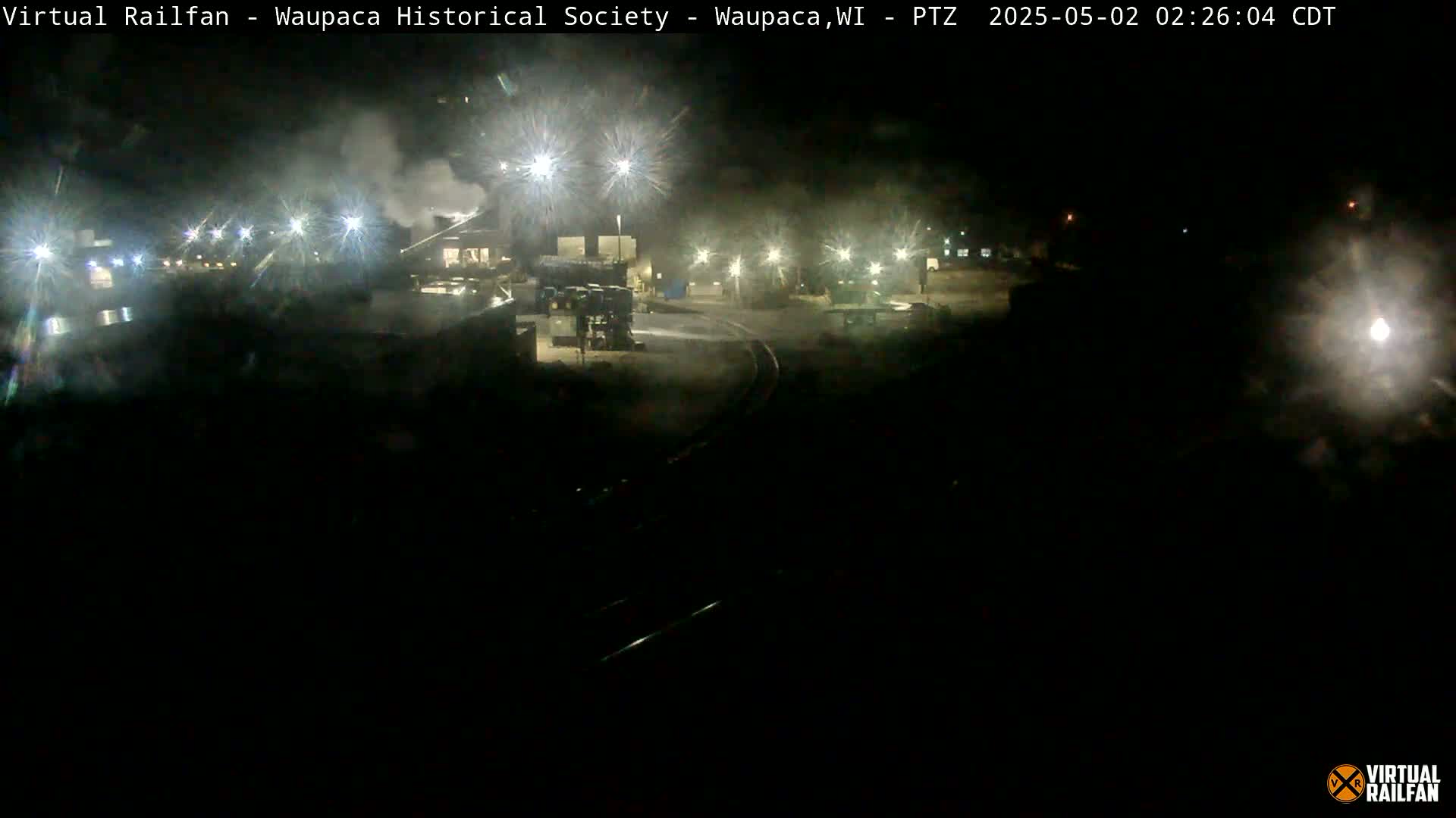 A nighttime, dimly lit industrial area is shown, featuring buildings, lights, and what appears to be a rail line.