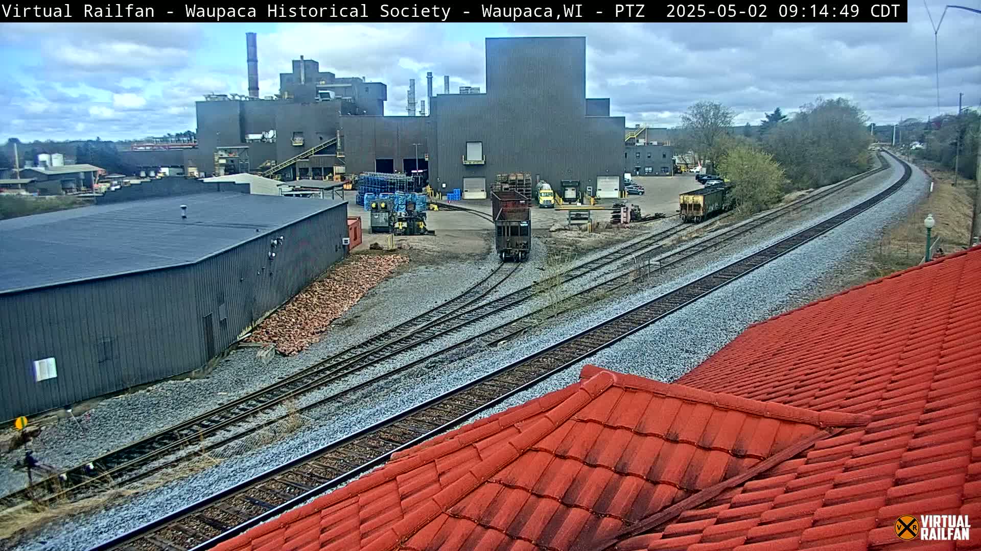 A large industrial building is situated near railroad tracks on an overcast day, with a rail car visible near a red tiled roof.