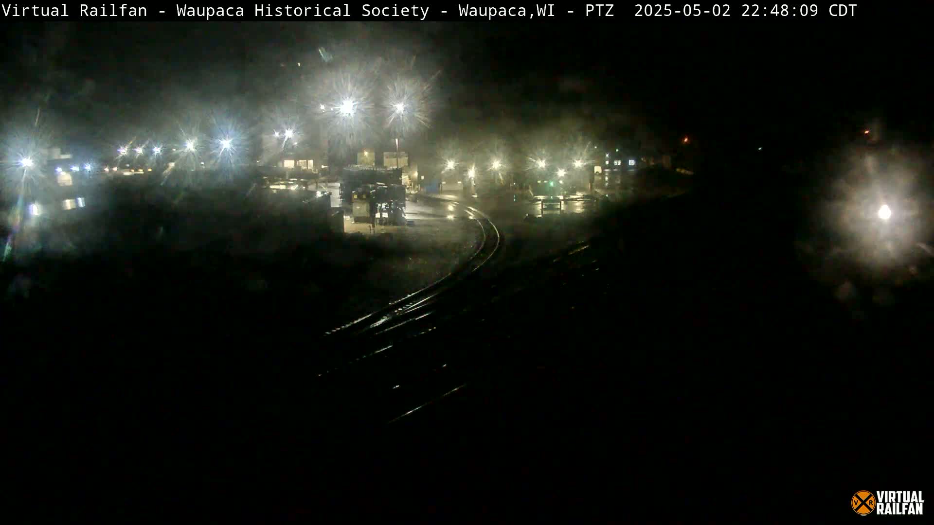 A nighttime, wet view of railroad tracks and a dimly lit industrial area with several bright lights.