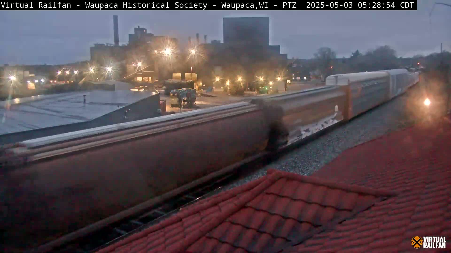 A long freight train passes a factory under a twilight sky.