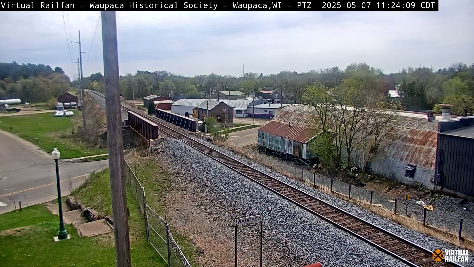 A mostly clear day reveals railroad tracks extending from a bridge over a road, passing near several buildings and trees.
