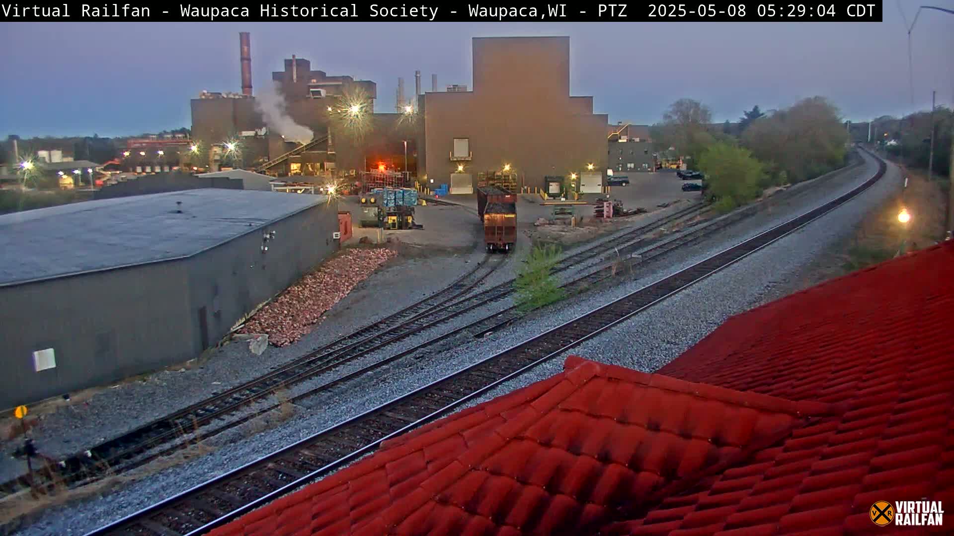 A train passes an industrial complex near a railway track under a twilight sky.