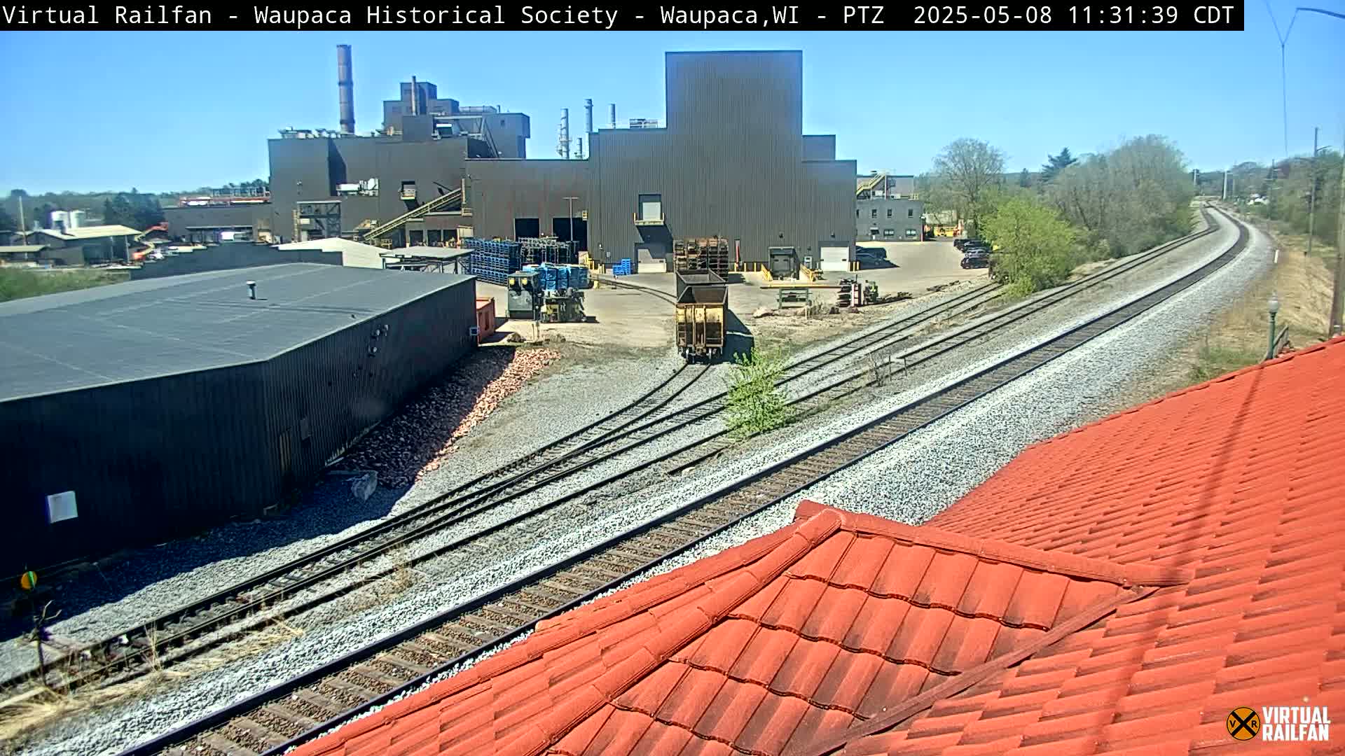 A rail yard with a large industrial building and a red-tiled roof in the foreground under sunny conditions.