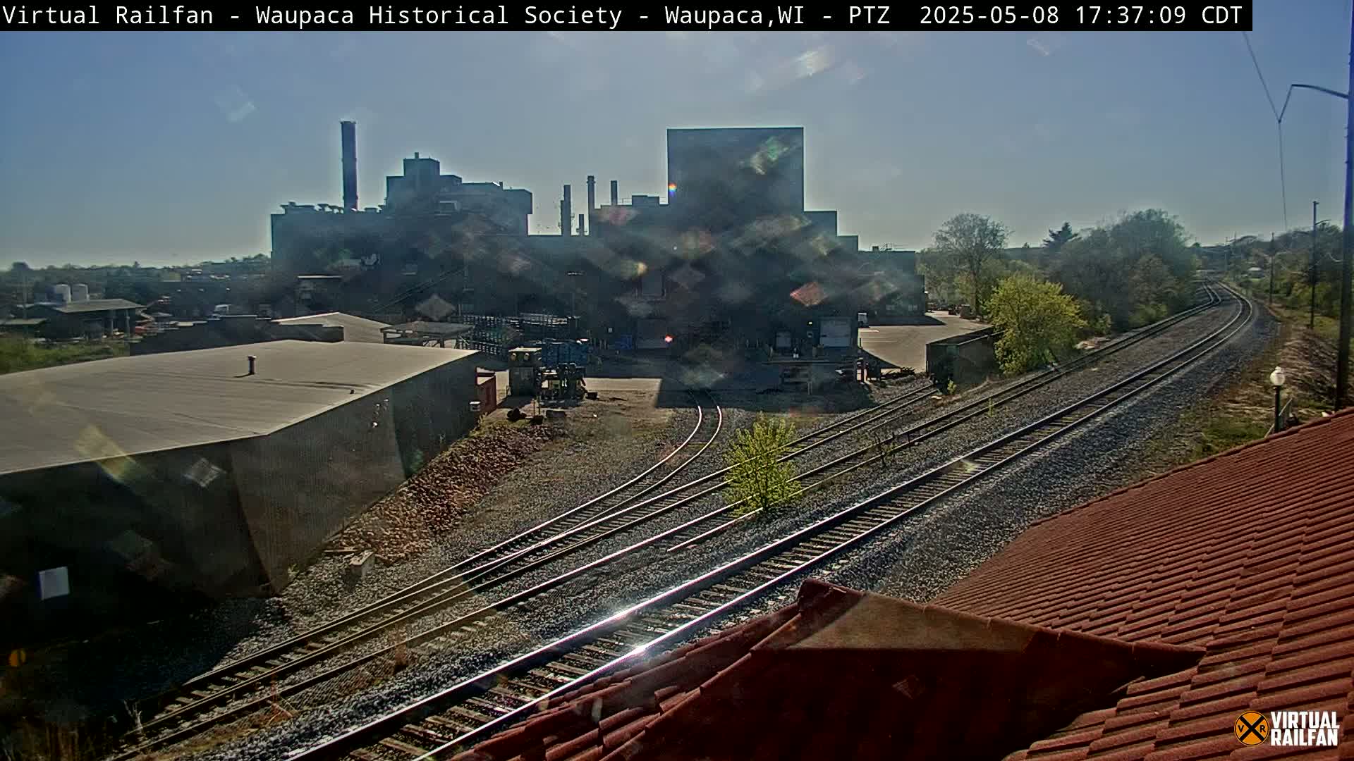 A large industrial building is visible in the distance beyond a network of train tracks on a sunny day.