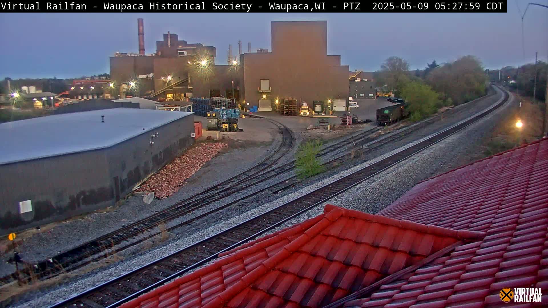 A dusk-lit industrial area with railroad tracks curving through it, and a red-tiled roof in the foreground.