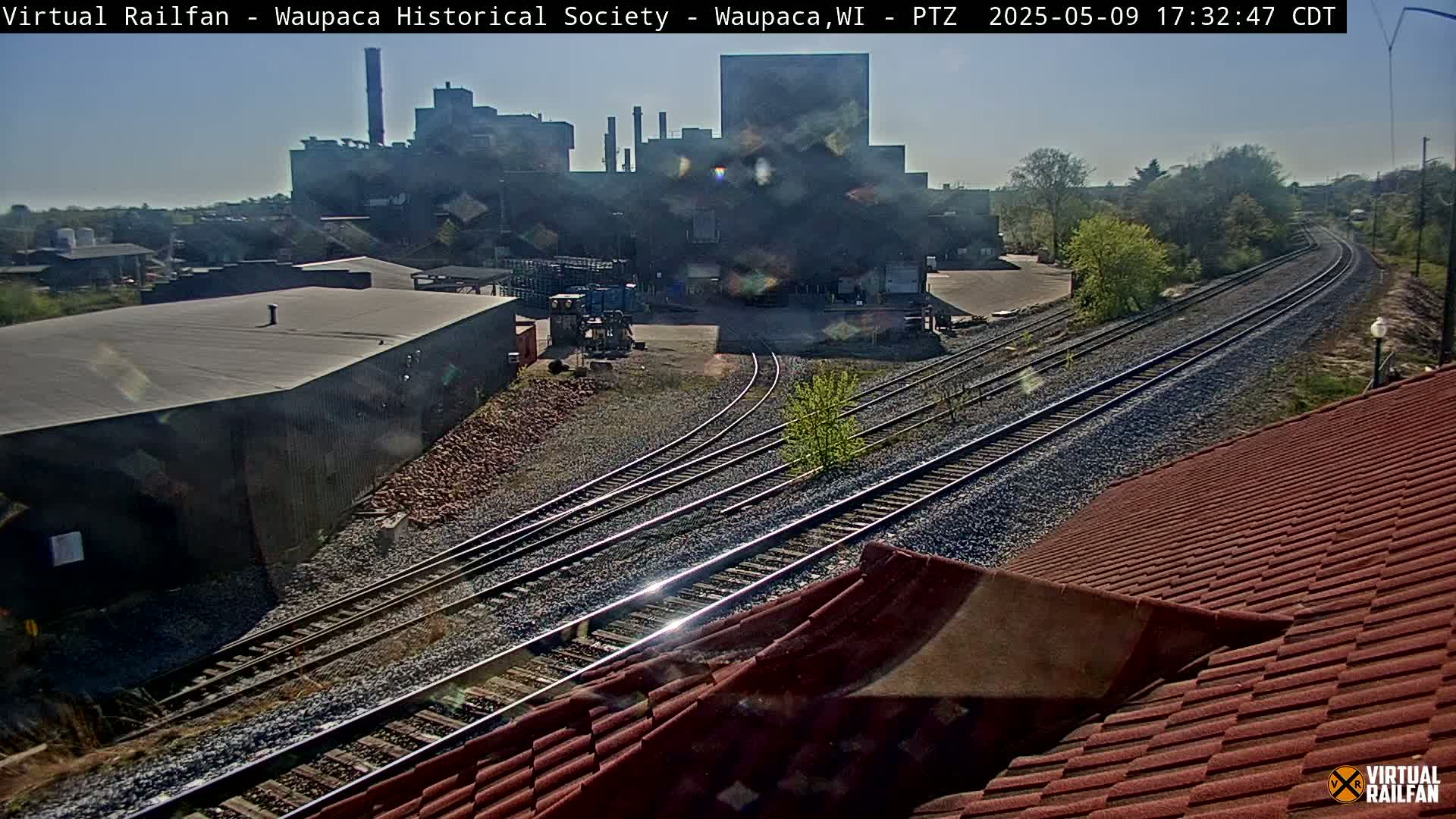 A large industrial building is visible beyond a railway with multiple tracks that diverge, under a sunny sky.