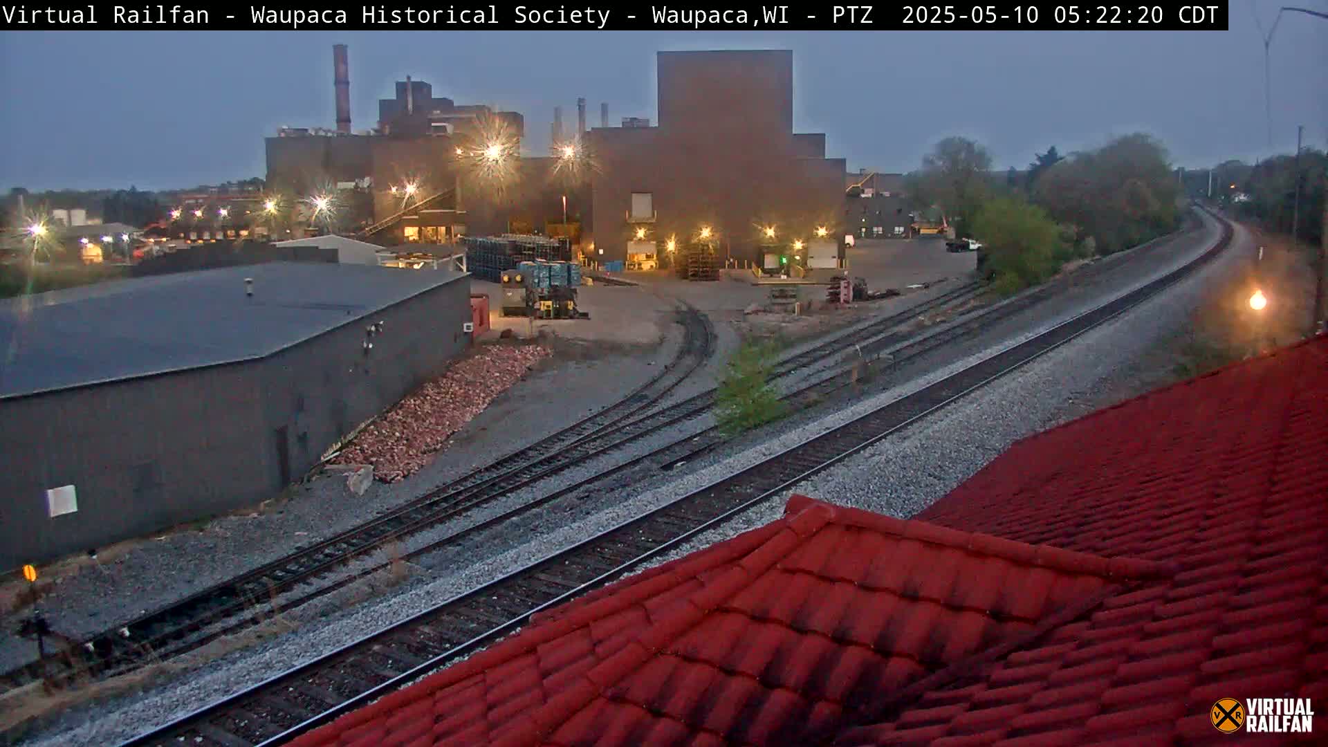 A nighttime view of a factory complex and railroad tracks, under a dusky sky.