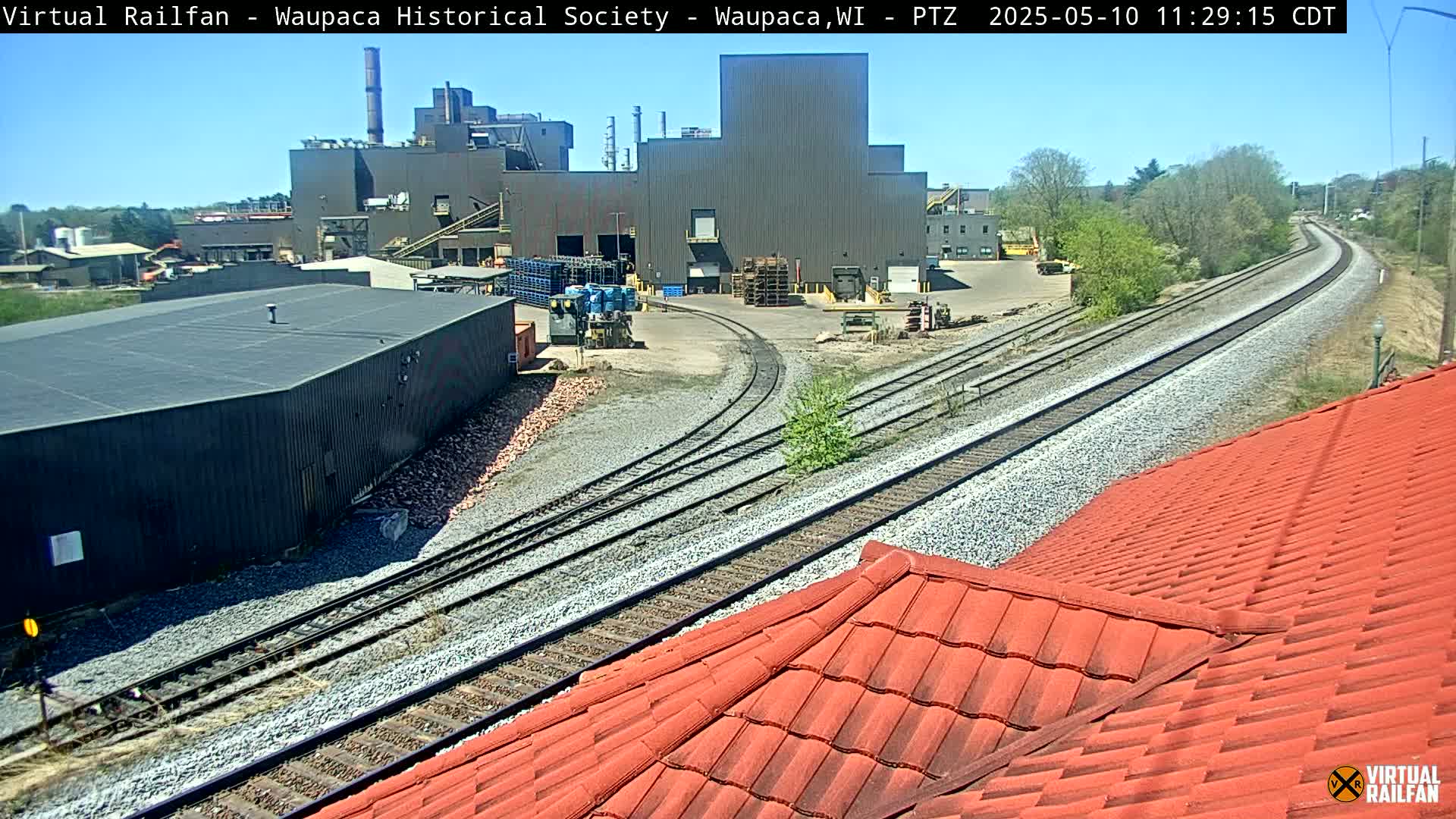 A sunny day shows a view of railroad tracks curving past an industrial building and a red-tiled roof.