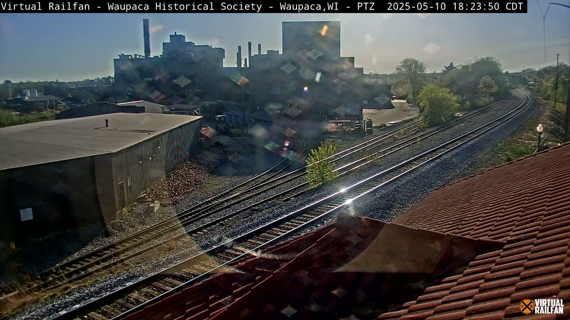 A view of railroad tracks intersecting near a large industrial building on a sunny day.