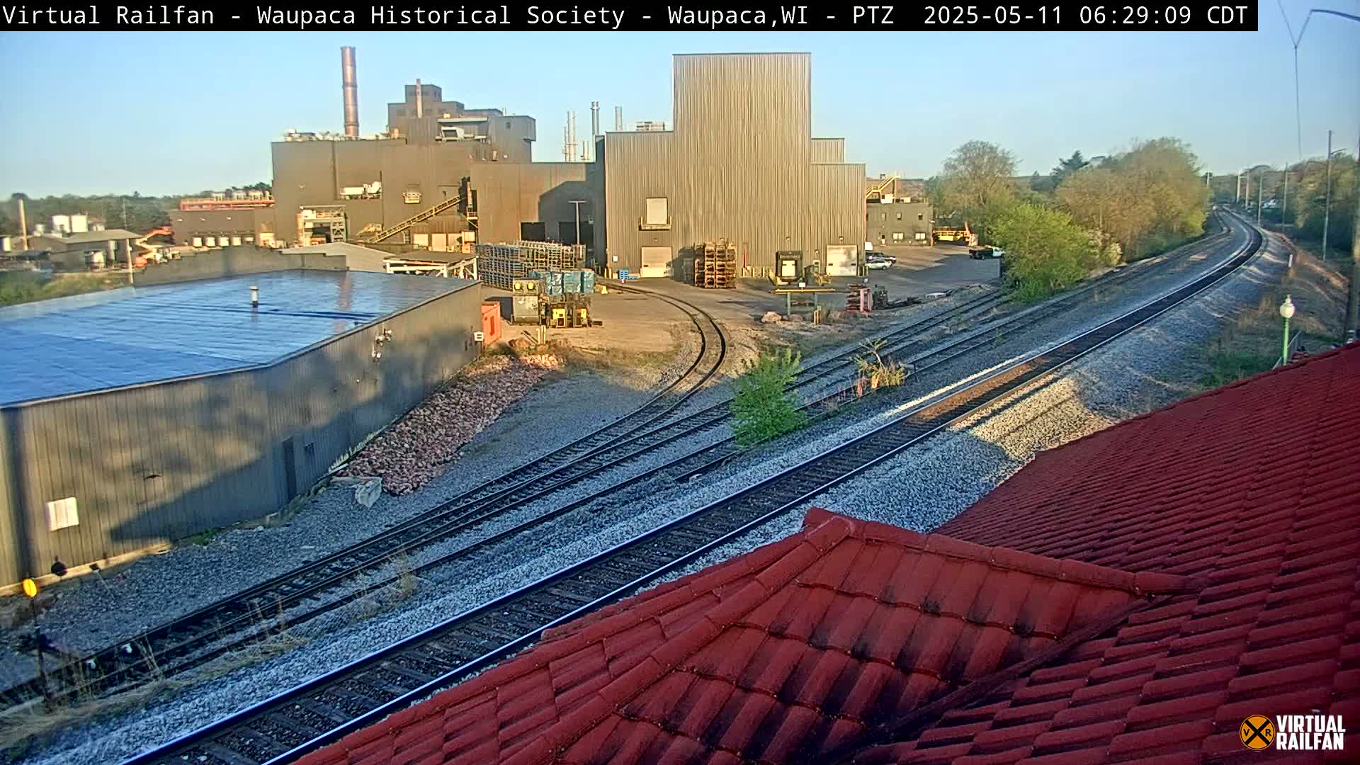 A large industrial complex is visible alongside train tracks on a sunny morning.