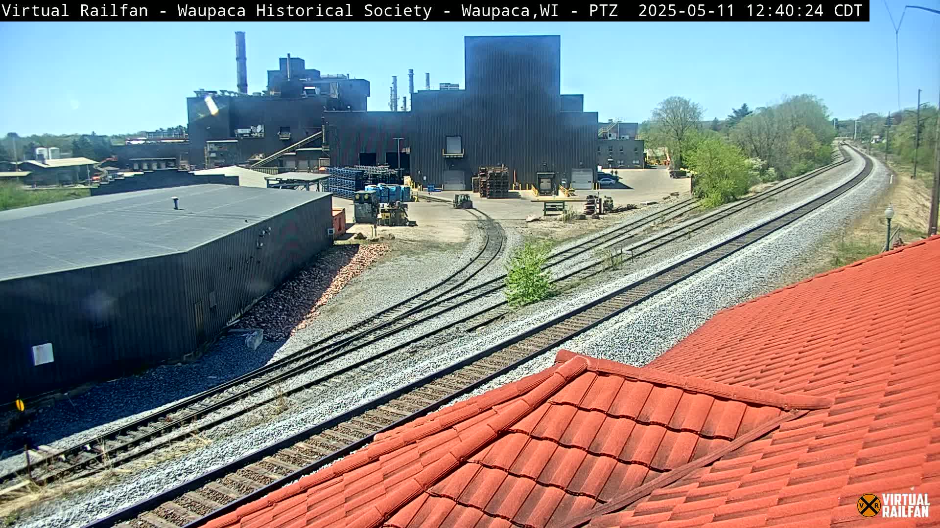 A large industrial complex is visible near a set of train tracks on a sunny day.