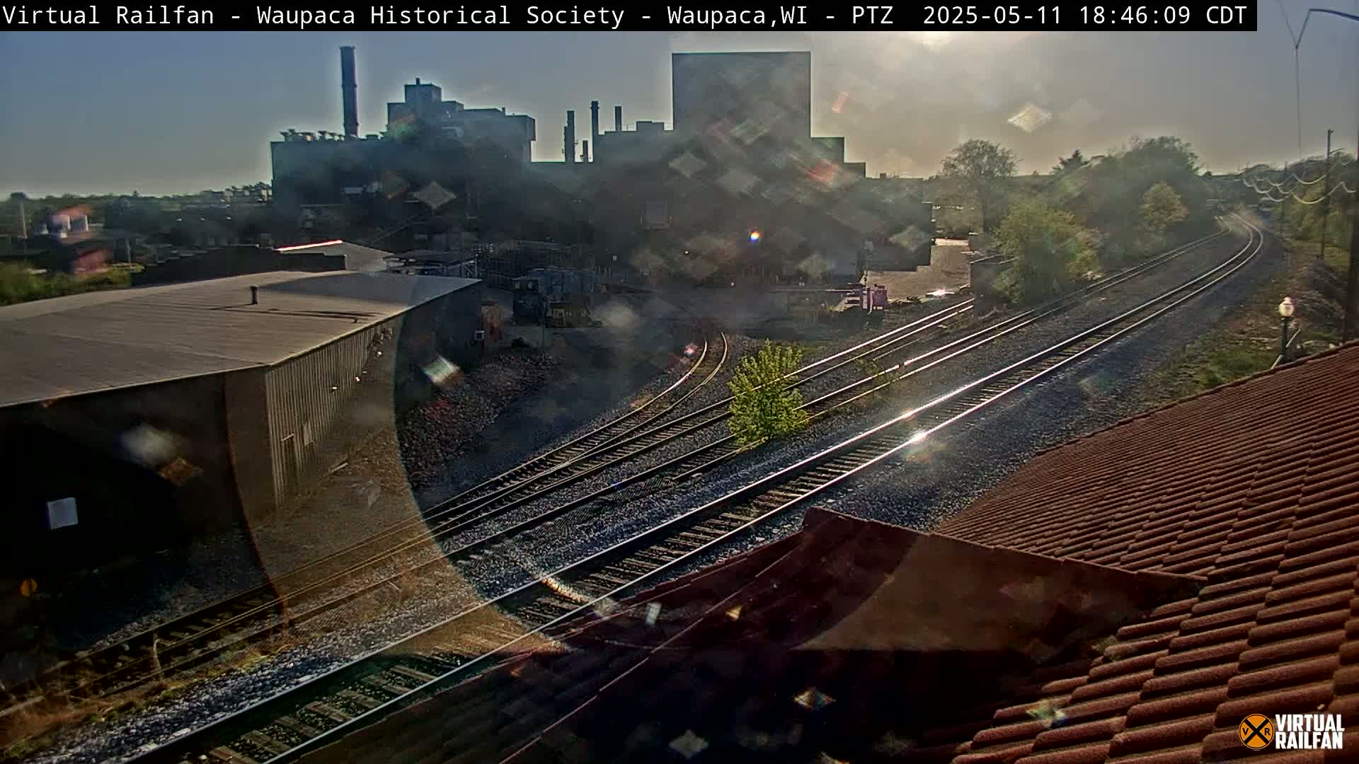 A high-angle, long shot shows railroad tracks curving away from a large industrial building under a partly sunny sky.