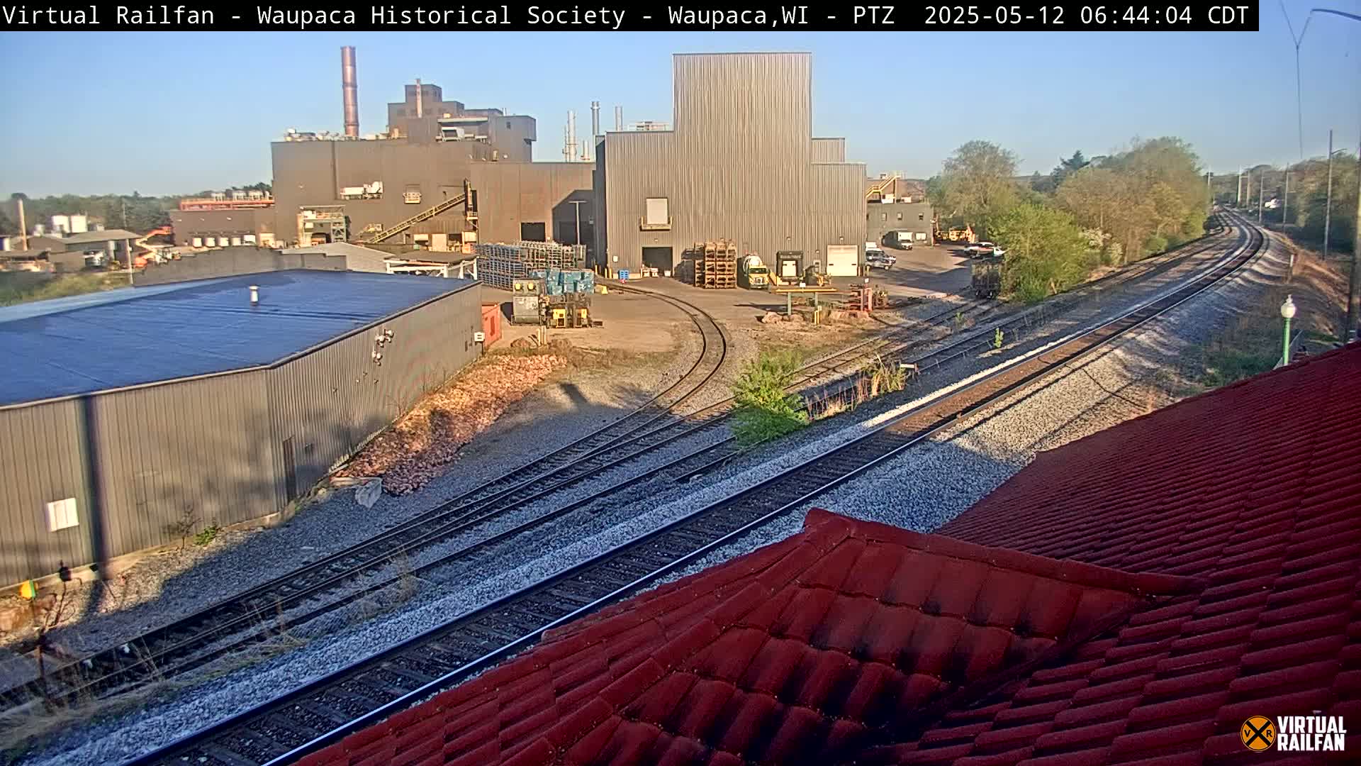 A sunny day reveals a large industrial complex near railroad tracks, with a red roofed structure in the foreground.