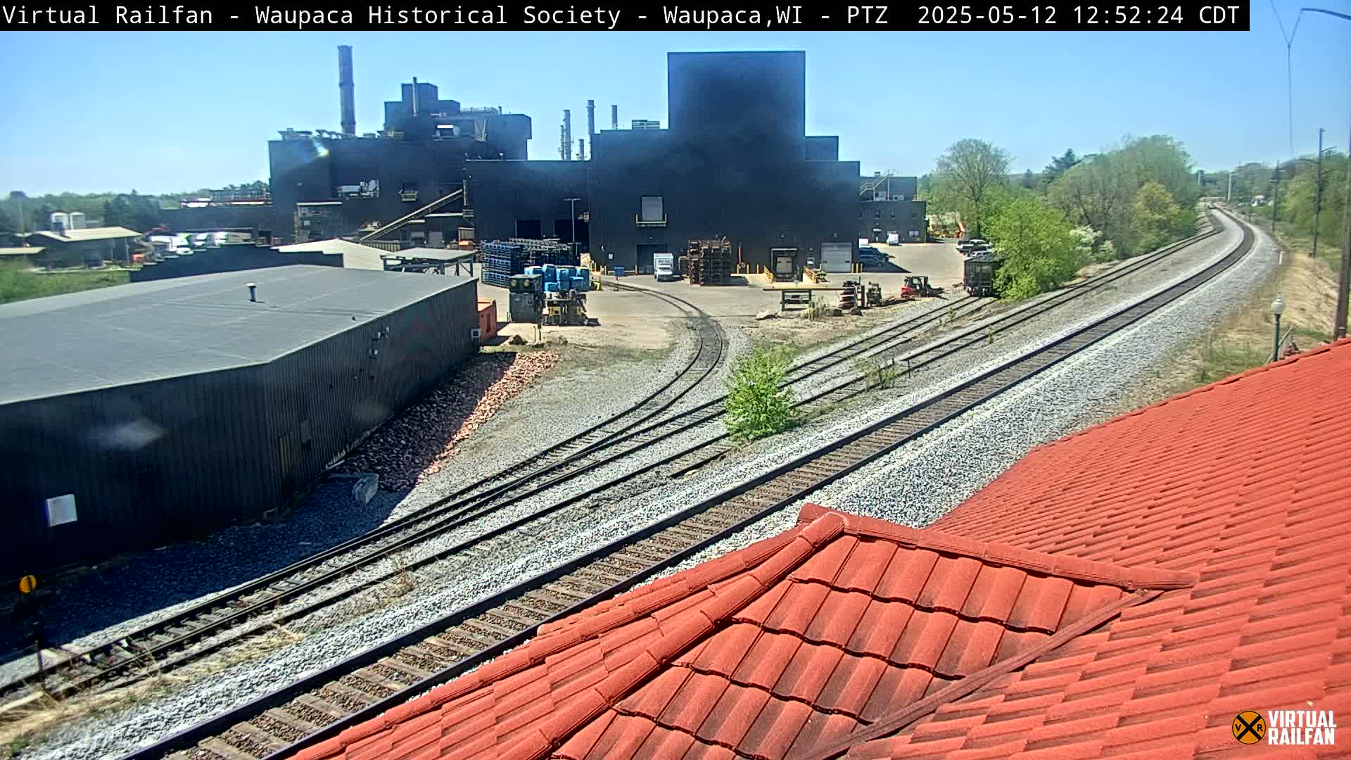 A large industrial building is visible beyond railroad tracks on a sunny day, partially obscured by a red tile roof in the foreground.