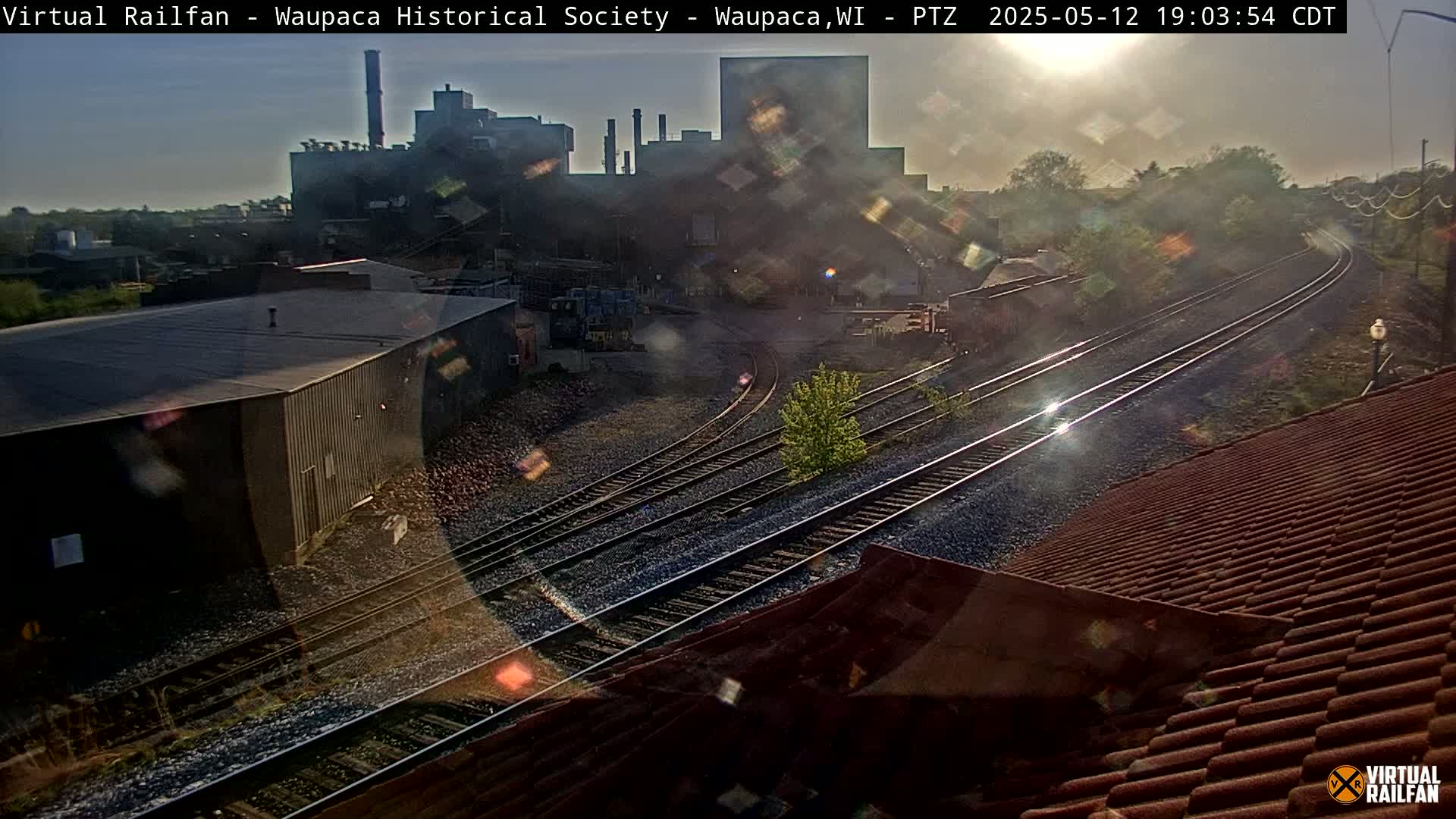 A hazy, sunlit view of railroad tracks curving past industrial buildings and a factory, with a red-tiled roof partially obscuring the foreground.