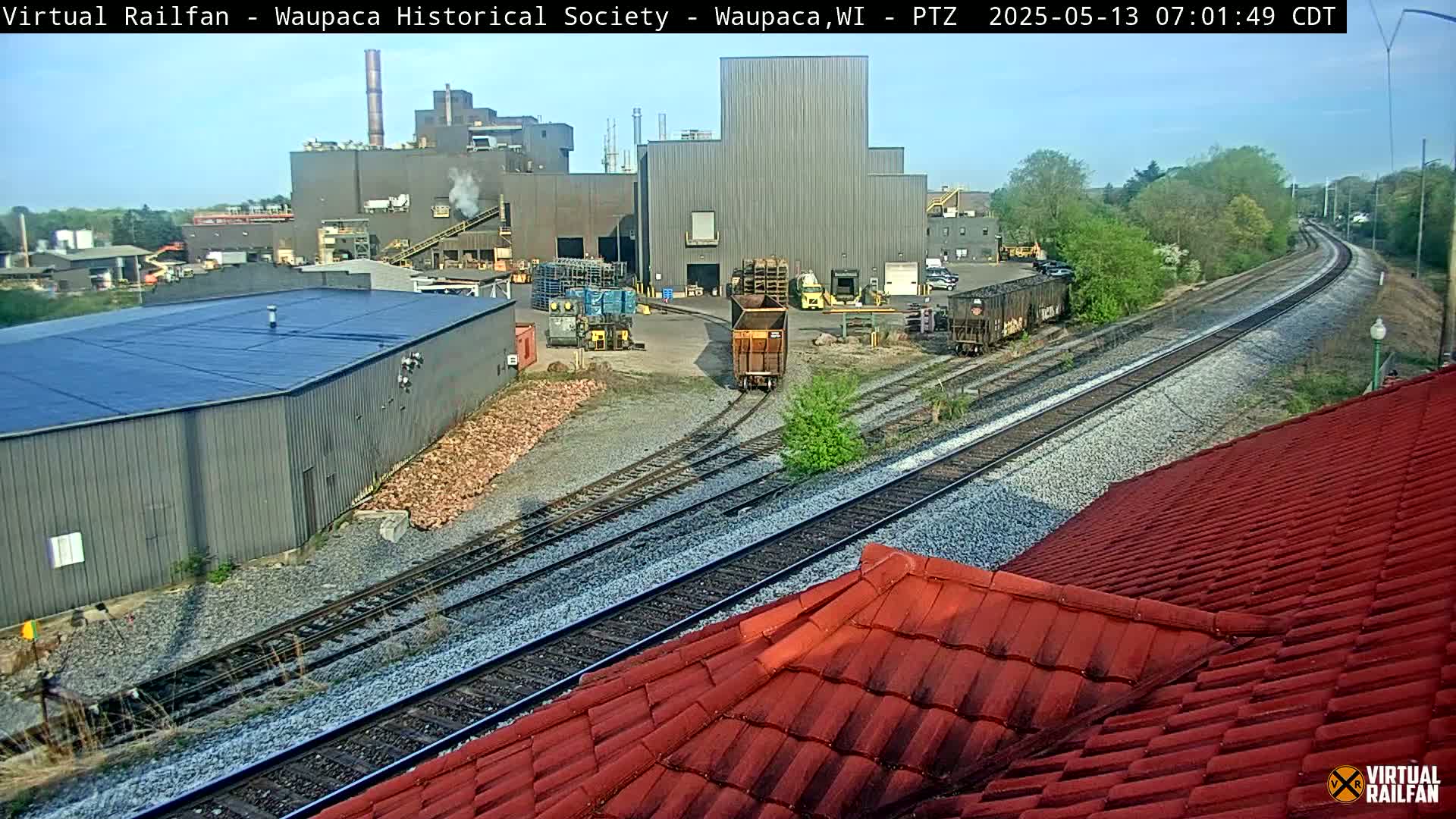 A factory complex is visible next to railroad tracks on a sunny day, with a red roof in the foreground.