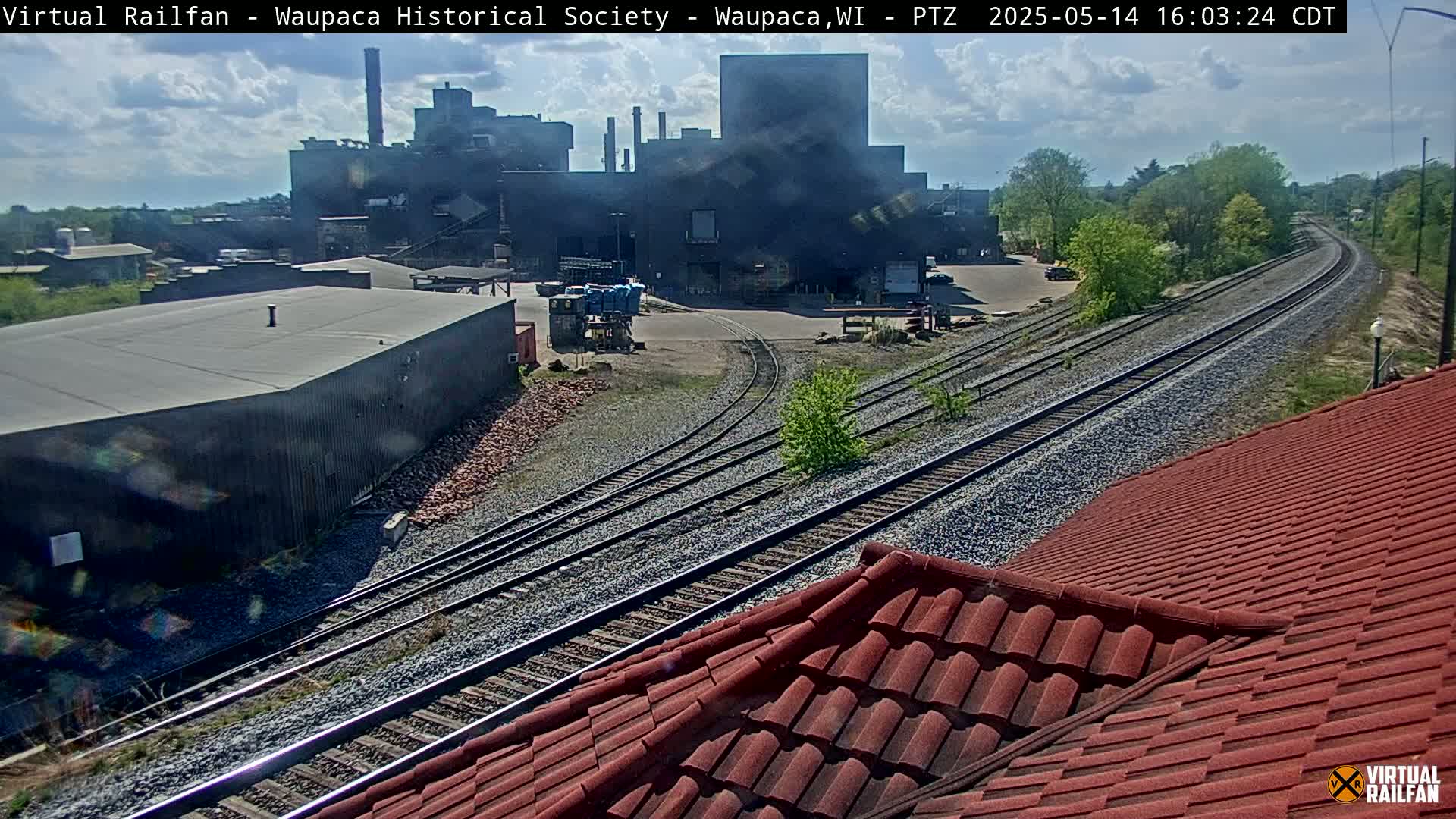 A view of railroad tracks leading to a large industrial building complex under a partly cloudy sky.