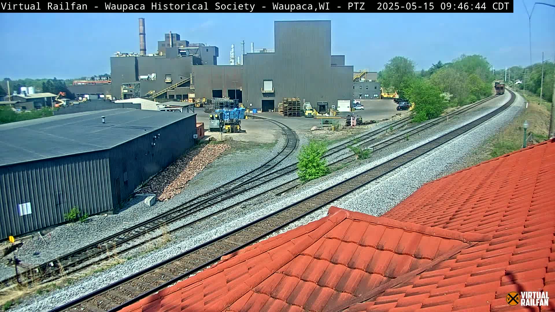 A large industrial building is situated beside railroad tracks on a sunny day, with a train visible in the distance and a red-tiled roof partially obscuring the foreground.