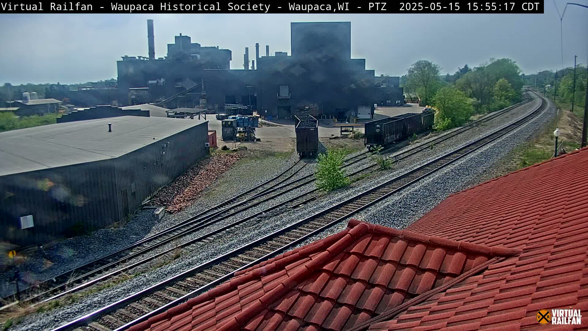 A view of railroad tracks leading to a large industrial building under a partly sunny sky.