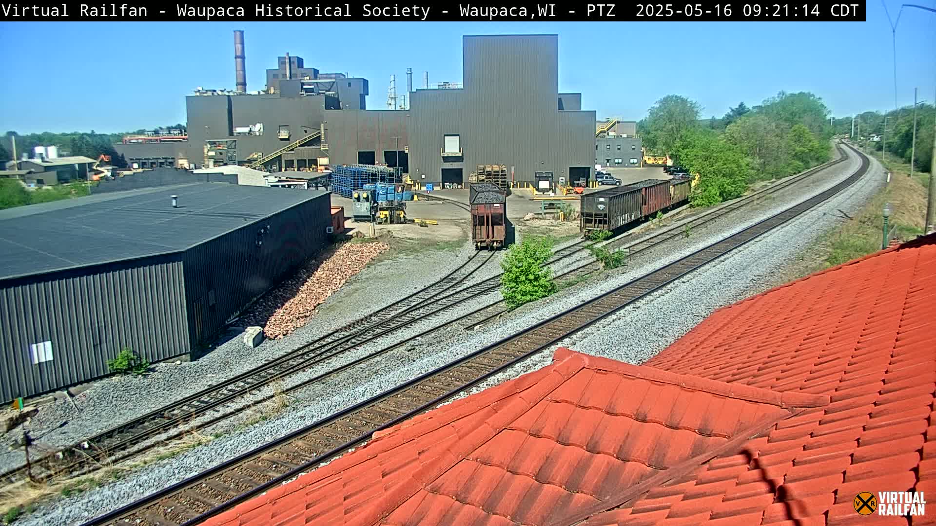 A section of railway lines snakes past an industrial complex under a clear, sunny sky, with a portion of a red tiled roof in the foreground.