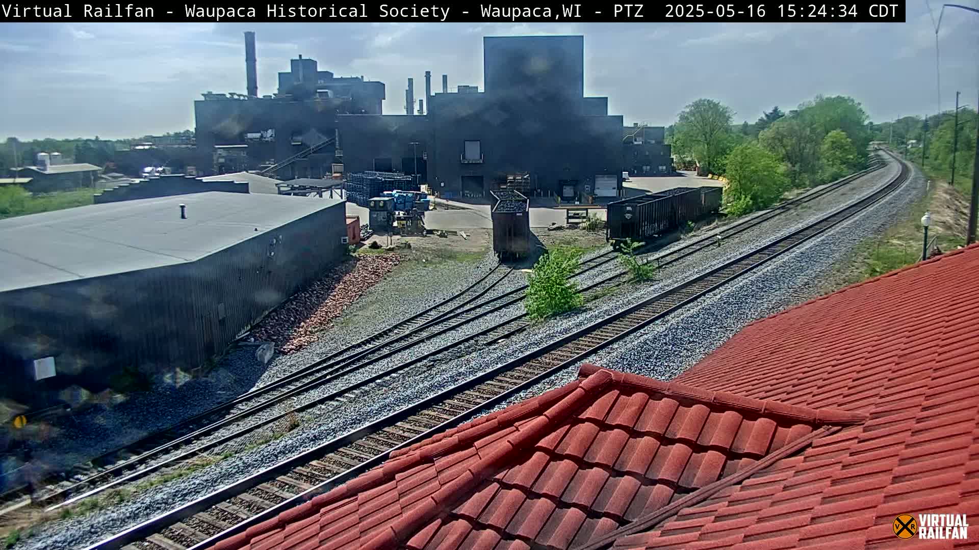 A mostly sunny day shows a large industrial building near railroad tracks with several rail cars, one of which is moving; a red roof is partially visible in the foreground.