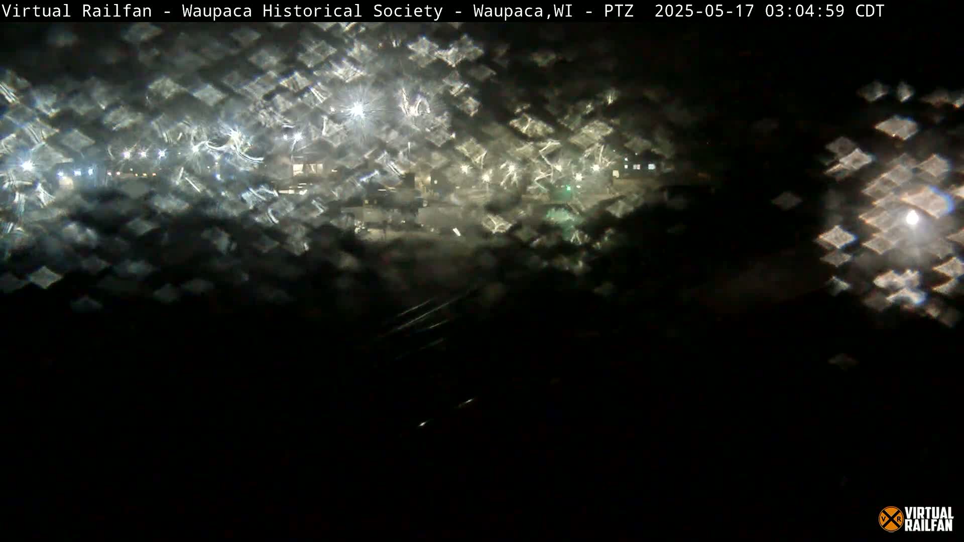 A nighttime view of a dimly lit area, seen through a rain-streaked window, shows scattered lights in the distance.