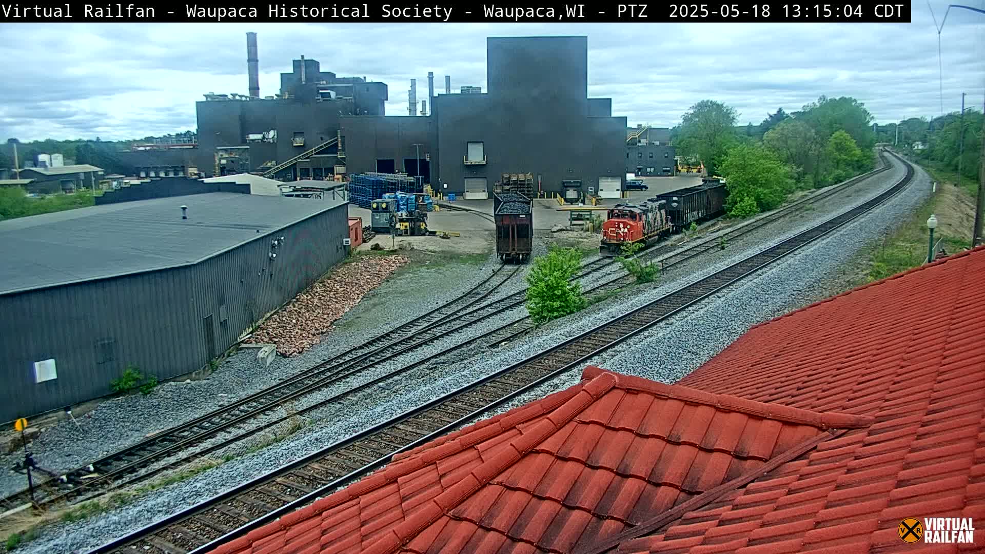 A large industrial building is visible beyond railroad tracks, with two locomotives and several railcars in the foreground, on an overcast day.