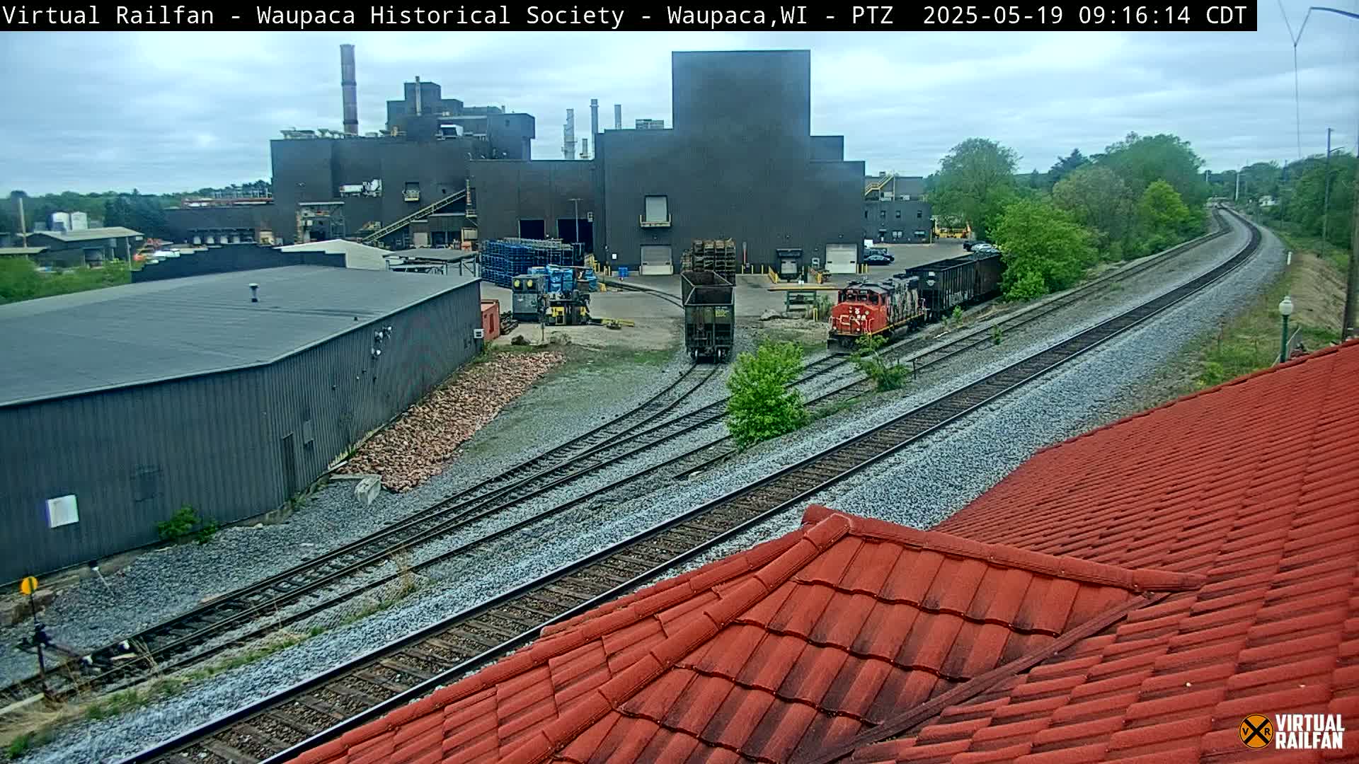 A large industrial building is seen beyond railroad tracks where two locomotives and a rail car are visible on an overcast day.