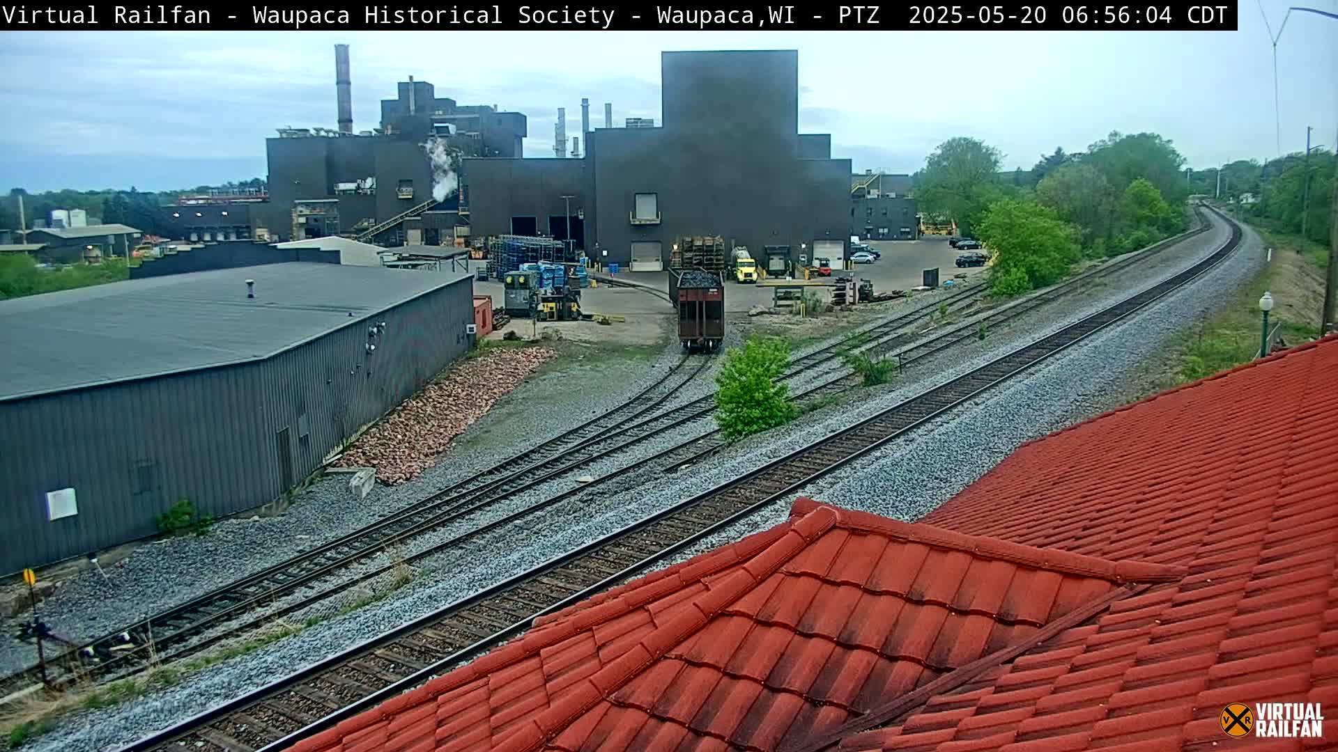 A railroad car approaches a large industrial building under an overcast sky, with railroad tracks extending into the distance.