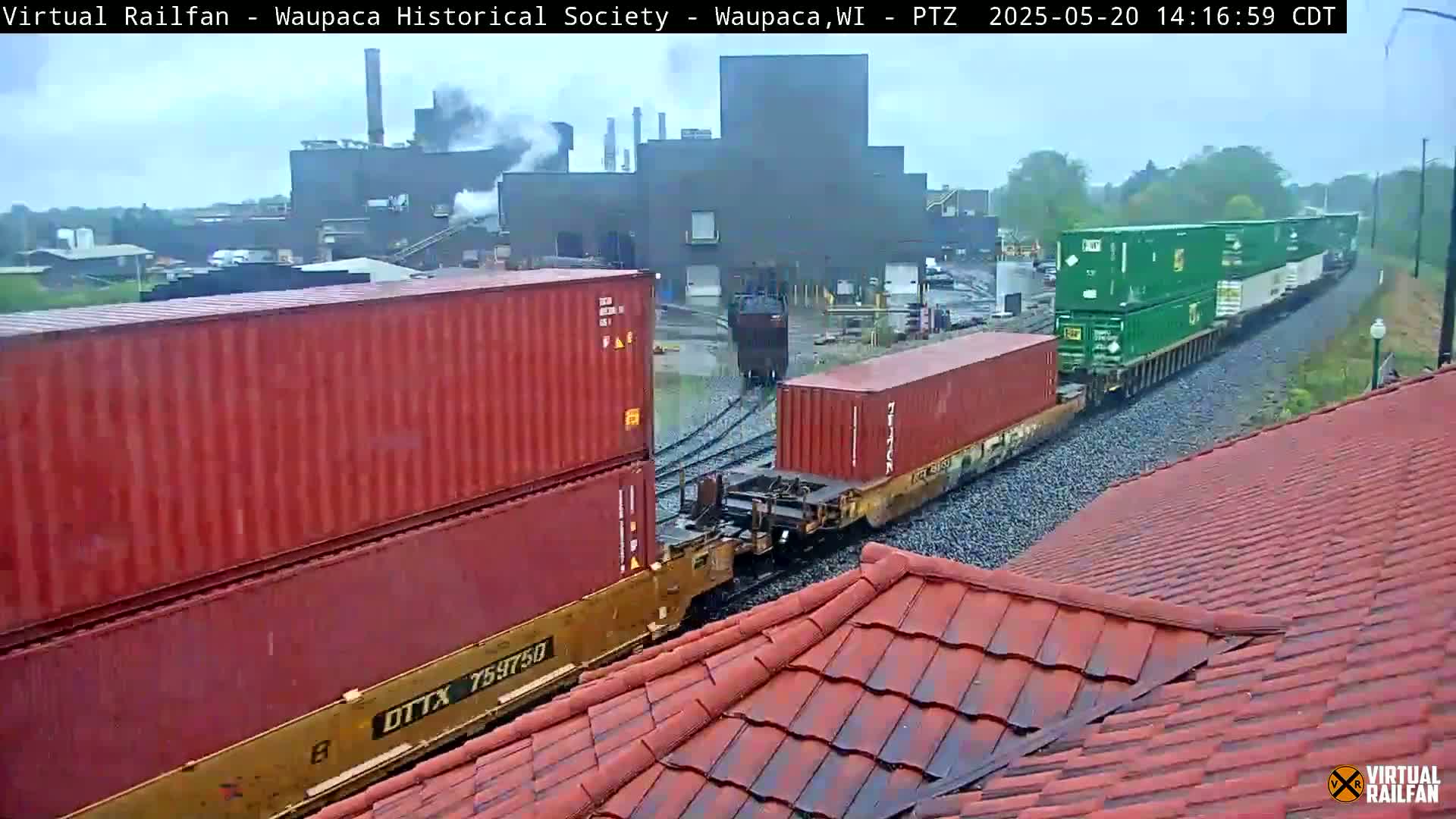 A freight train carrying stacked shipping containers passes a factory under an overcast sky.