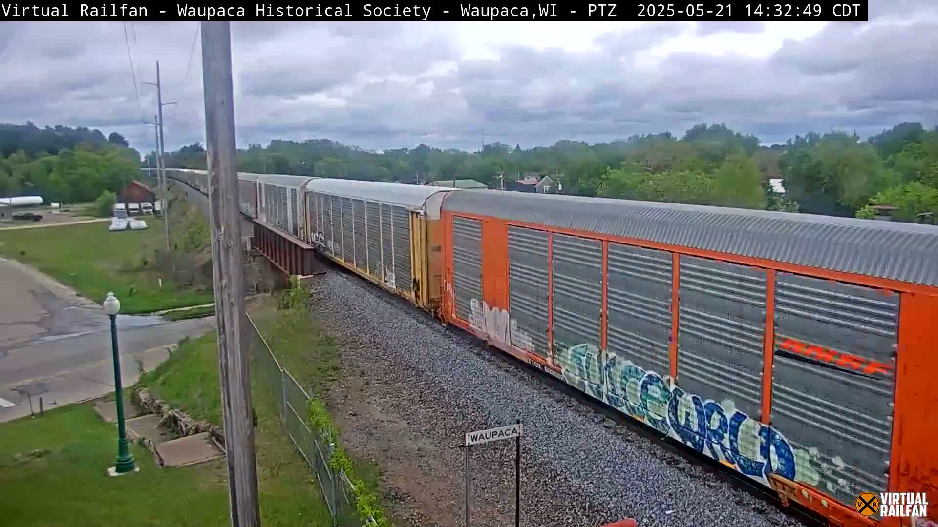 A long freight train with graffiti on its orange cars passes over a railway bridge under a partly cloudy sky.