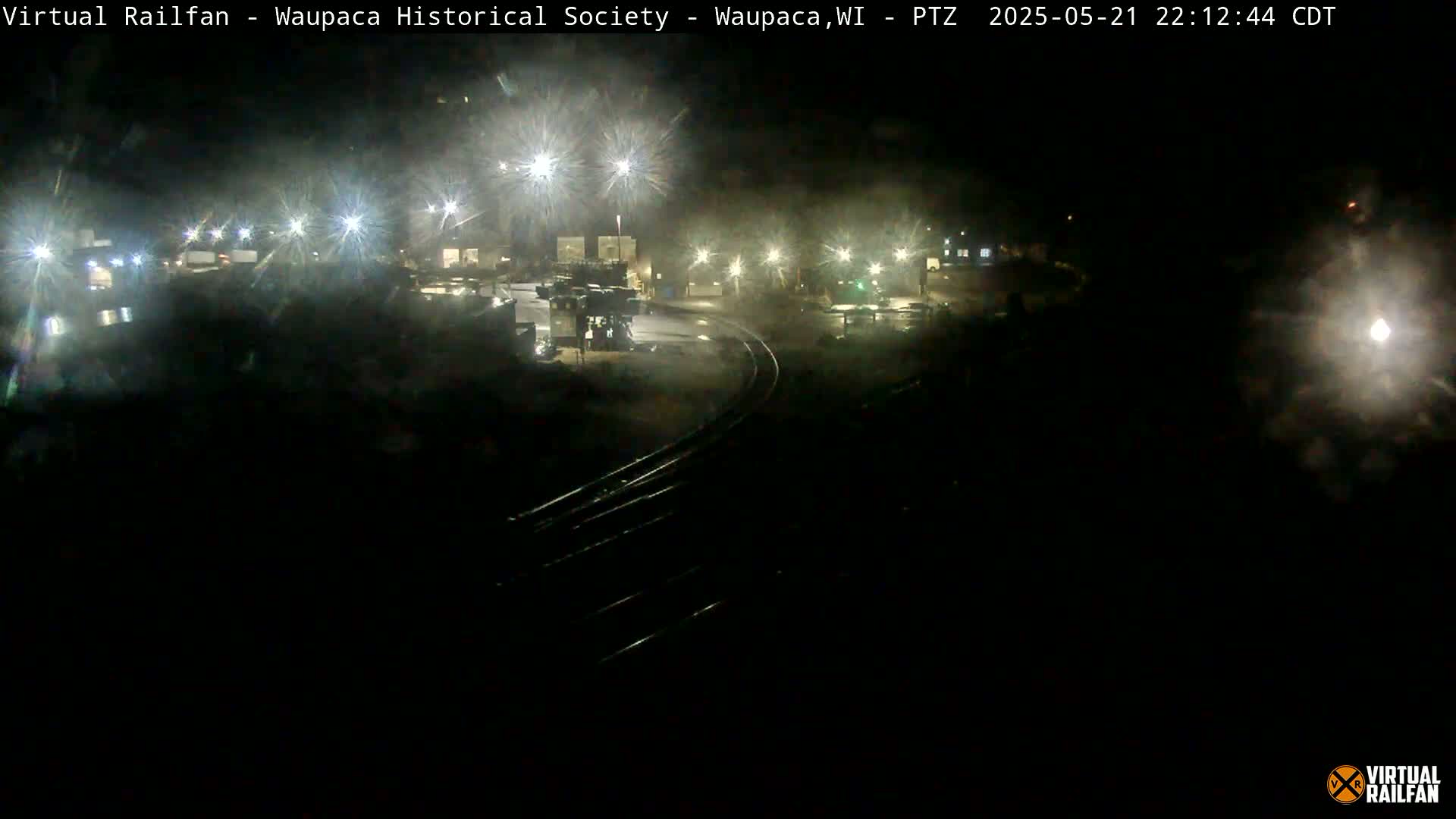 A nighttime, dimly lit view of a rail yard with some machinery and buildings visible under artificial lights.