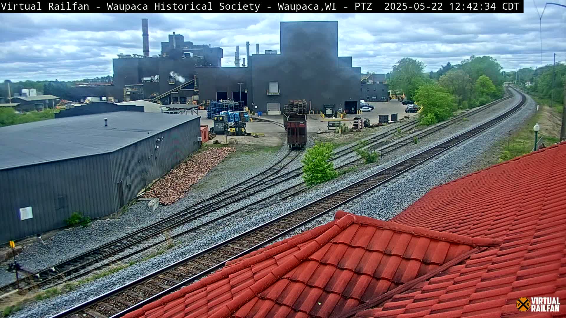 A railway runs alongside a large industrial building complex under a partly cloudy sky; a red-tiled roof is in the foreground.