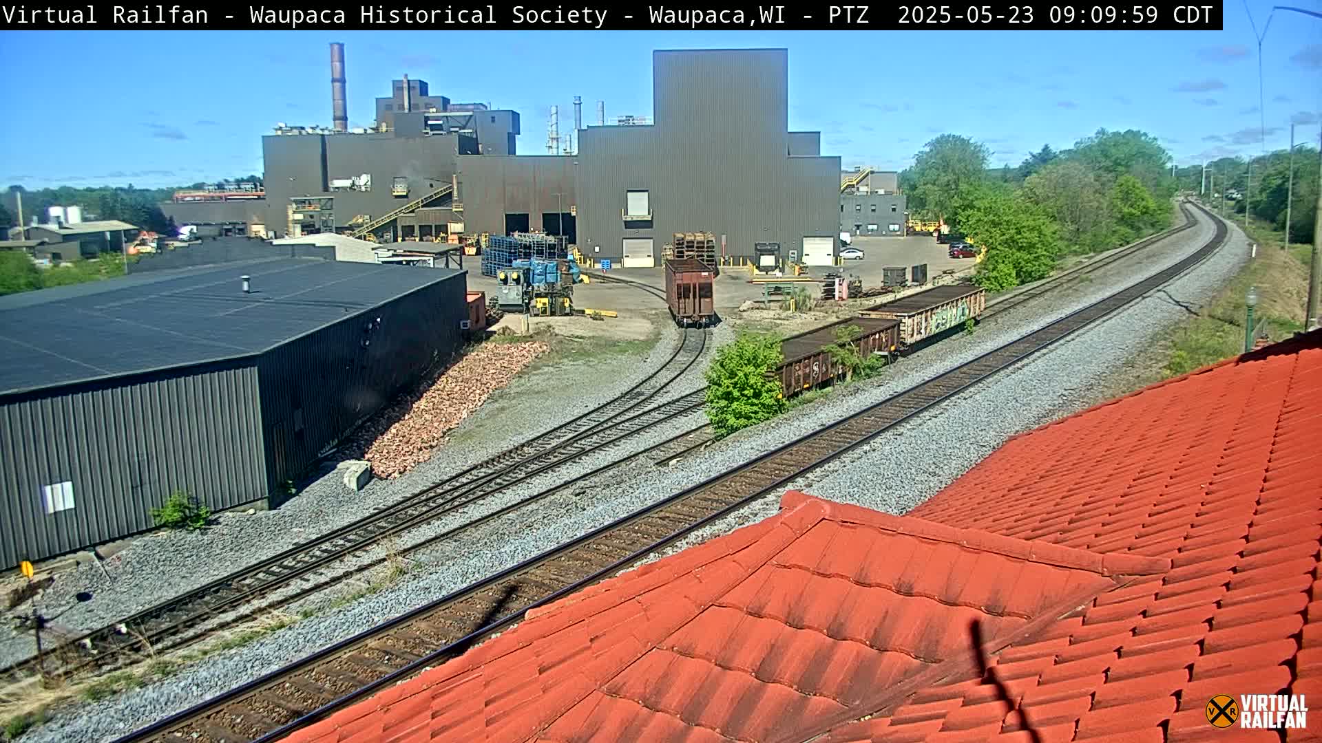 A train passes by an industrial complex on a sunny day, with railroad tracks extending into the distance, partially obscured by a red tiled roof in the foreground.