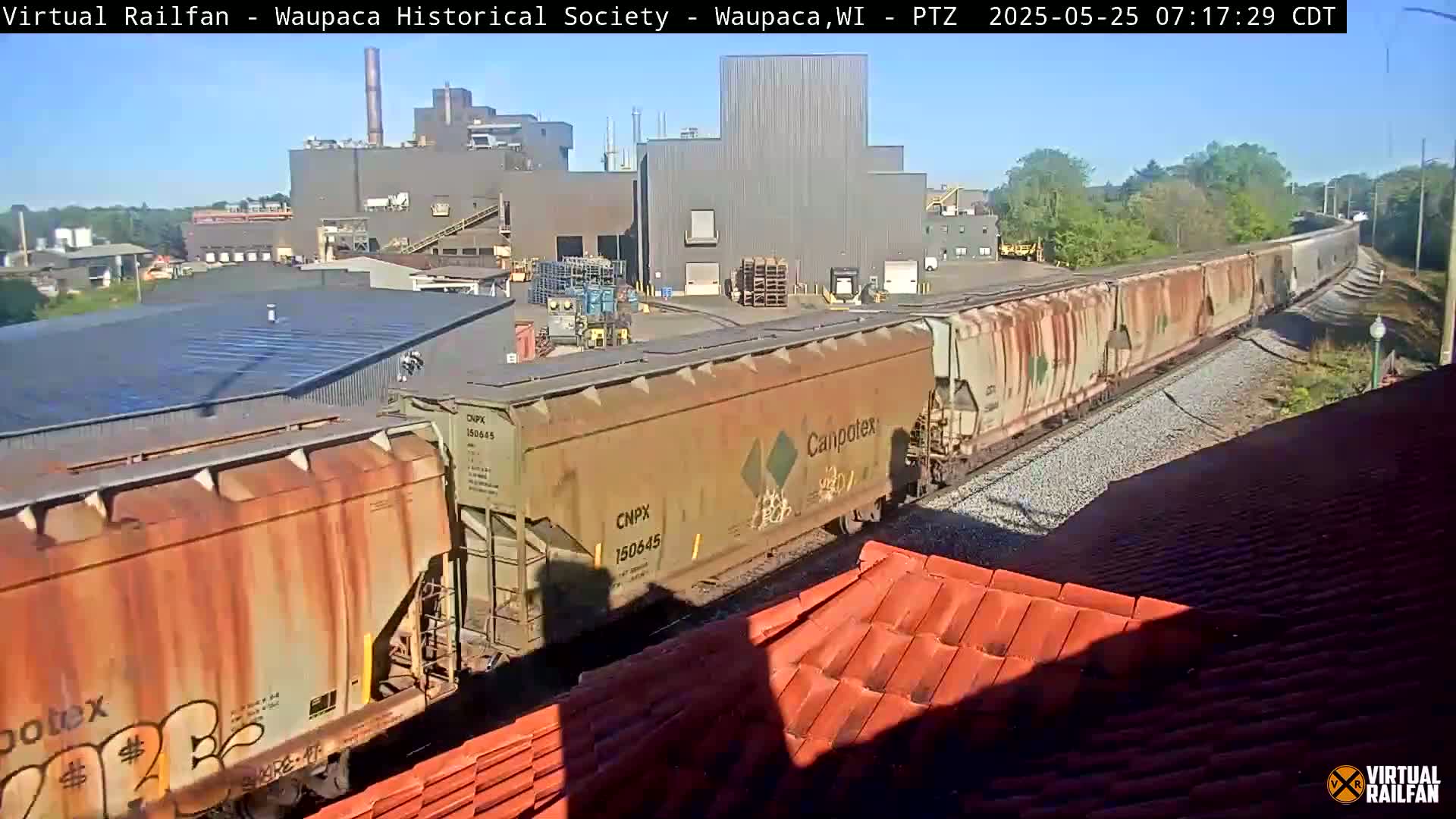 A long freight train passes by a large industrial building on a sunny day.