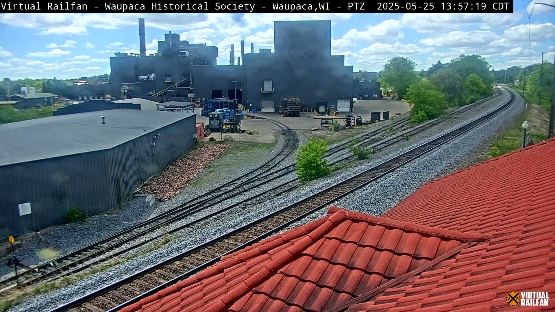 A view of train tracks curving past an industrial complex under a partly cloudy sky.