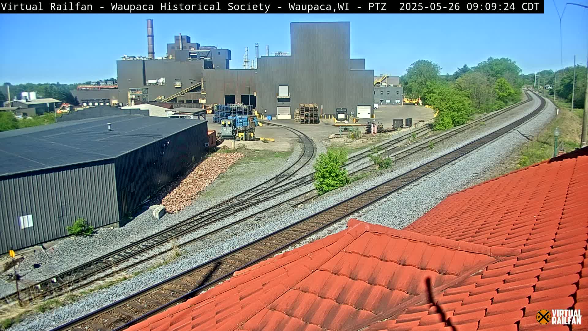 A view of industrial buildings and railway tracks under a sunny sky.