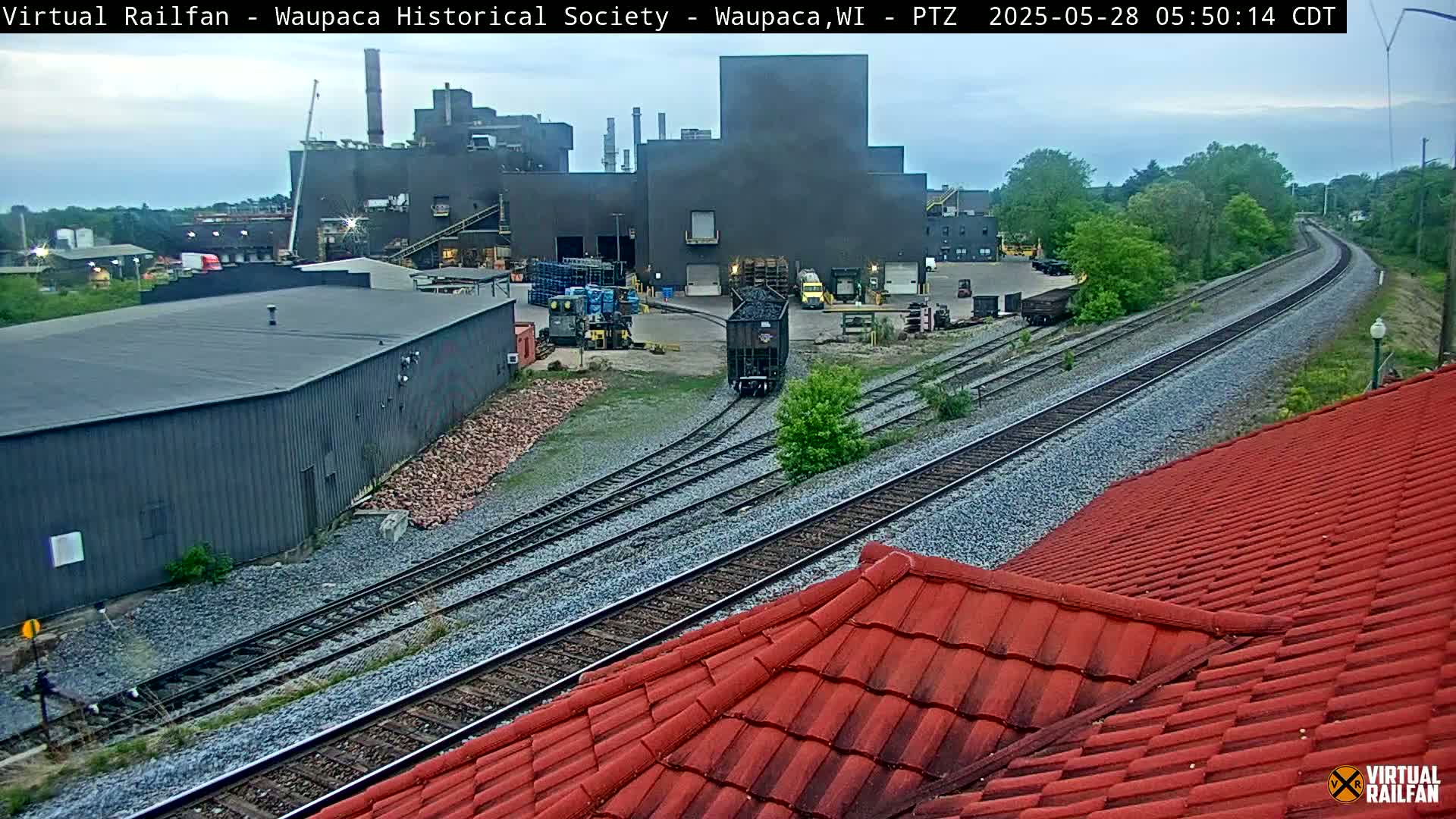 A dark-colored industrial building is situated near railroad tracks on an overcast day, with a single rail car visible on the tracks and a red roof in the foreground.