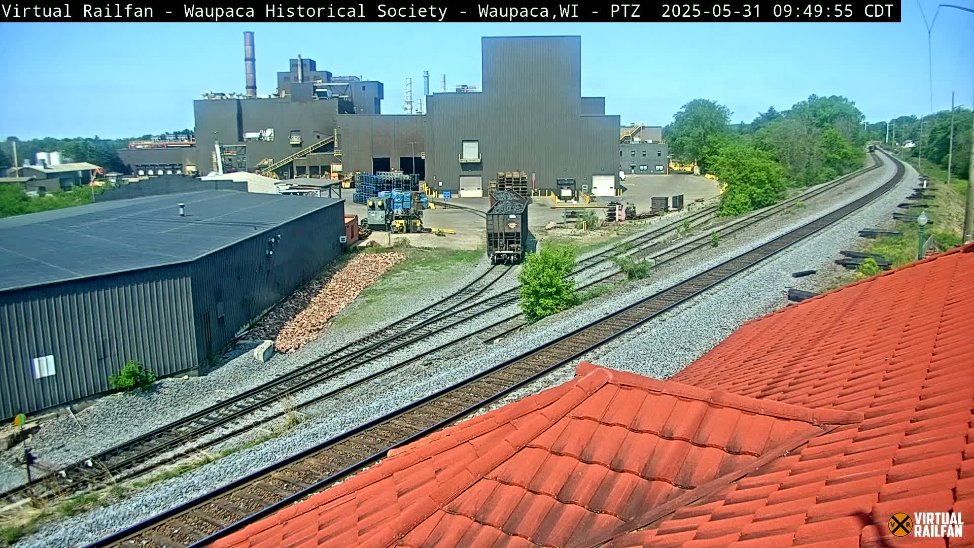 A train car moves along railway tracks toward a large industrial building under a clear blue sky.