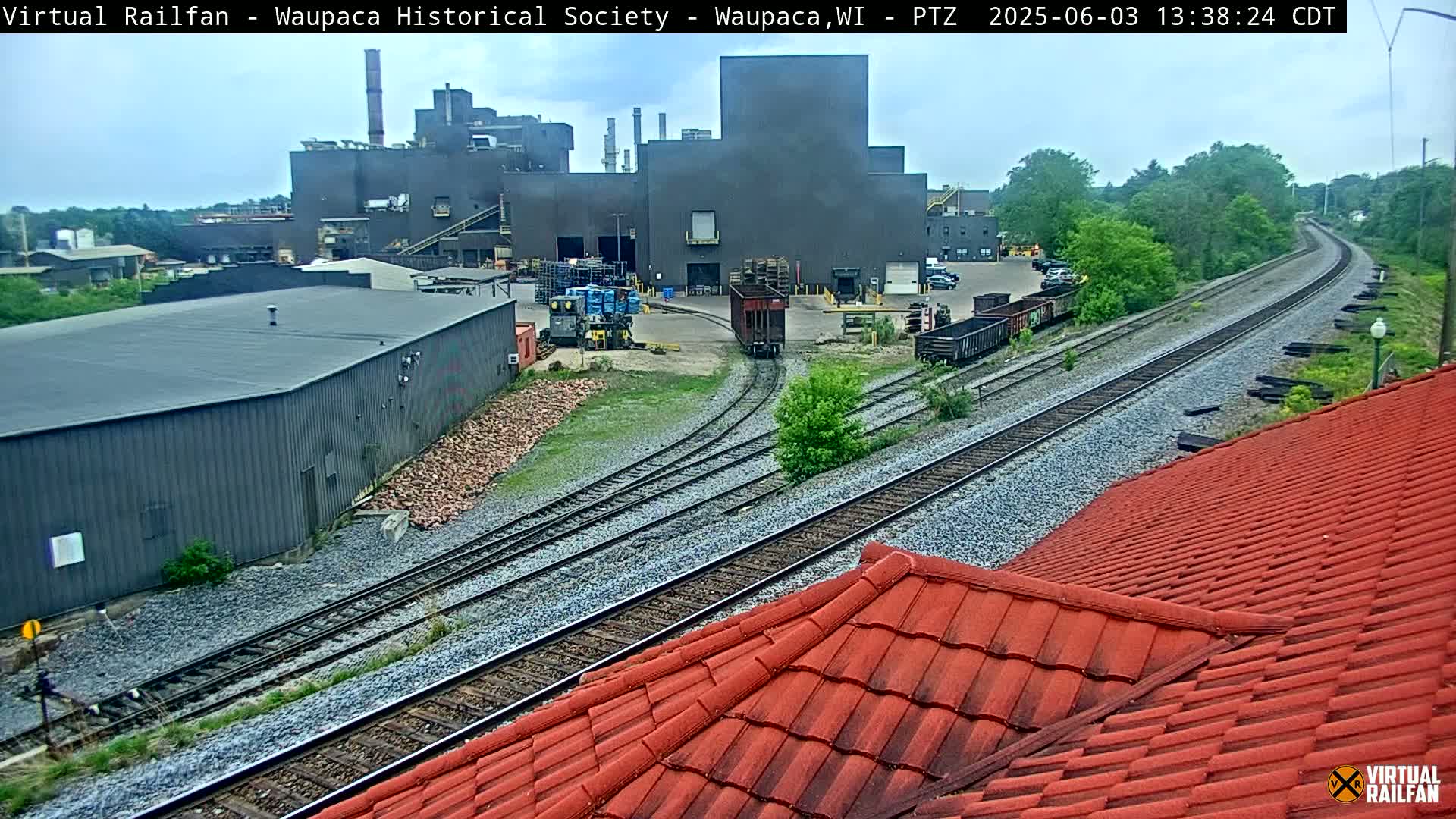A large industrial building is visible next to railroad tracks on an overcast day, with several rail cars near the building and a red-tiled roof in the foreground.