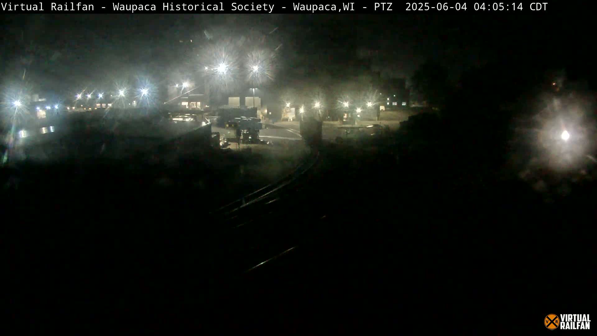 A nighttime view of a dimly lit industrial area with several buildings and vehicles, under cloudy conditions.